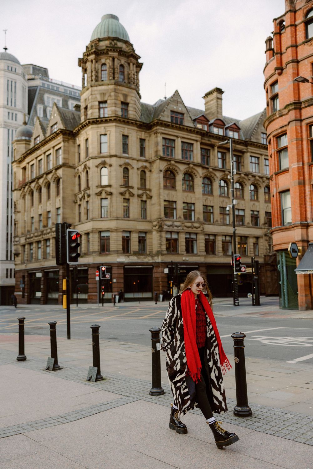 A person in a red coat and patterned dress walks past historic Victorian architecture in Manchester city center.