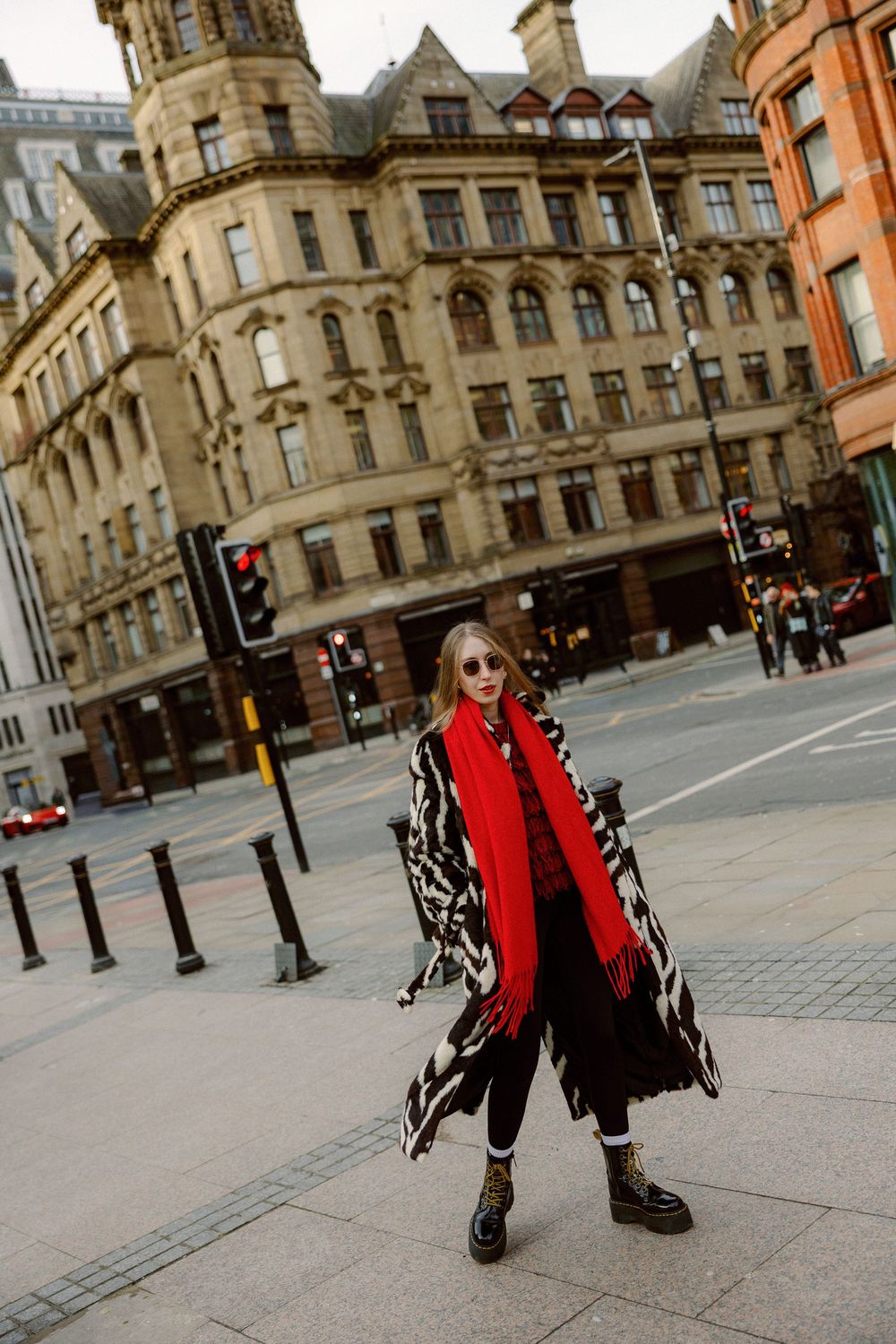 Person in a bright red jacket and black pants poses on a historic European city street with ornate architecture.
