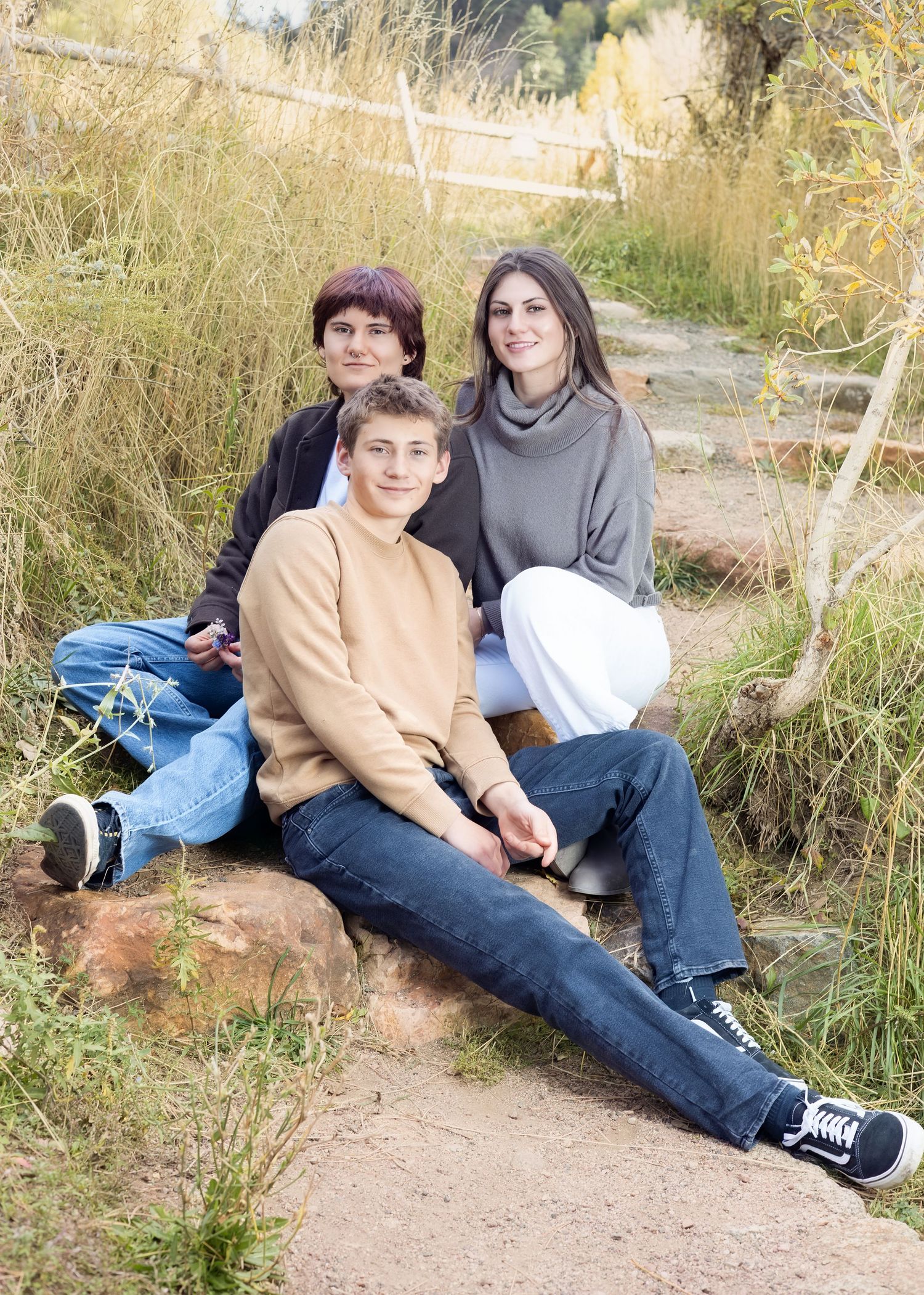 Three people sit together on a dirt path surrounded by tall grass and autumn foliage on a sunny day.