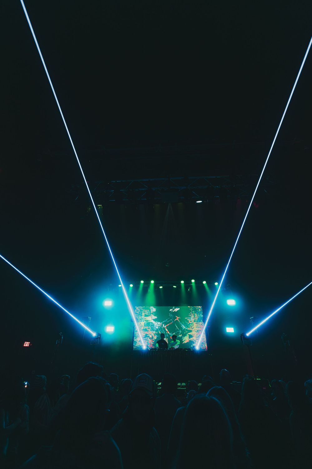 Blue laser beams illuminate a dark concert stage with green accent lighting creating a dramatic performance atmosphere.