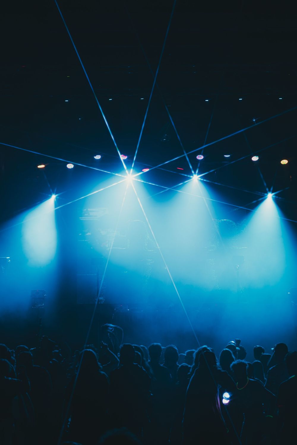 Concert venue illuminated with dramatic blue stage lights and laser beams shining over silhouetted crowd.