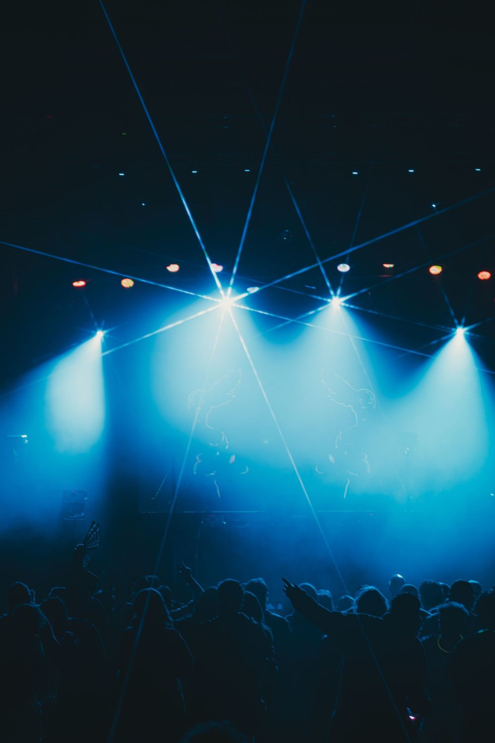 Concert crowd silhouettes under dramatic blue stage lighting with bright beams cutting through atmospheric haze.