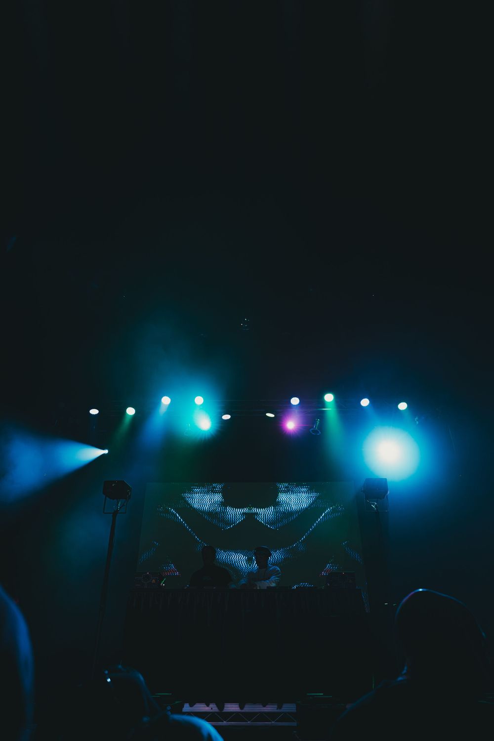 Silhouetted stage equipment and lighting rigs create a dramatic atmosphere with blue and purple lights in a dark venue.