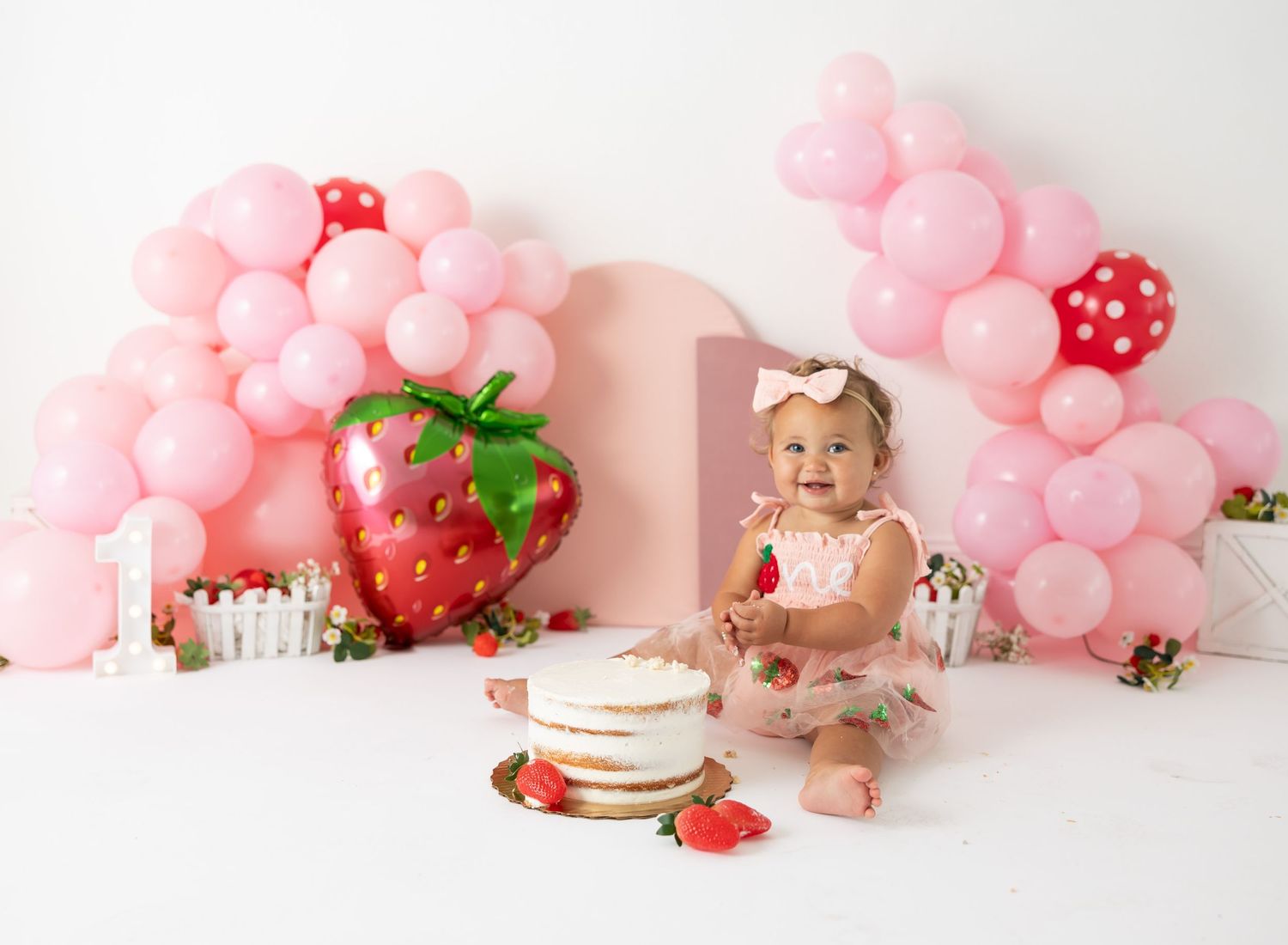 Baby celebrating birthday with strawberry themed decor and pink balloon arch.