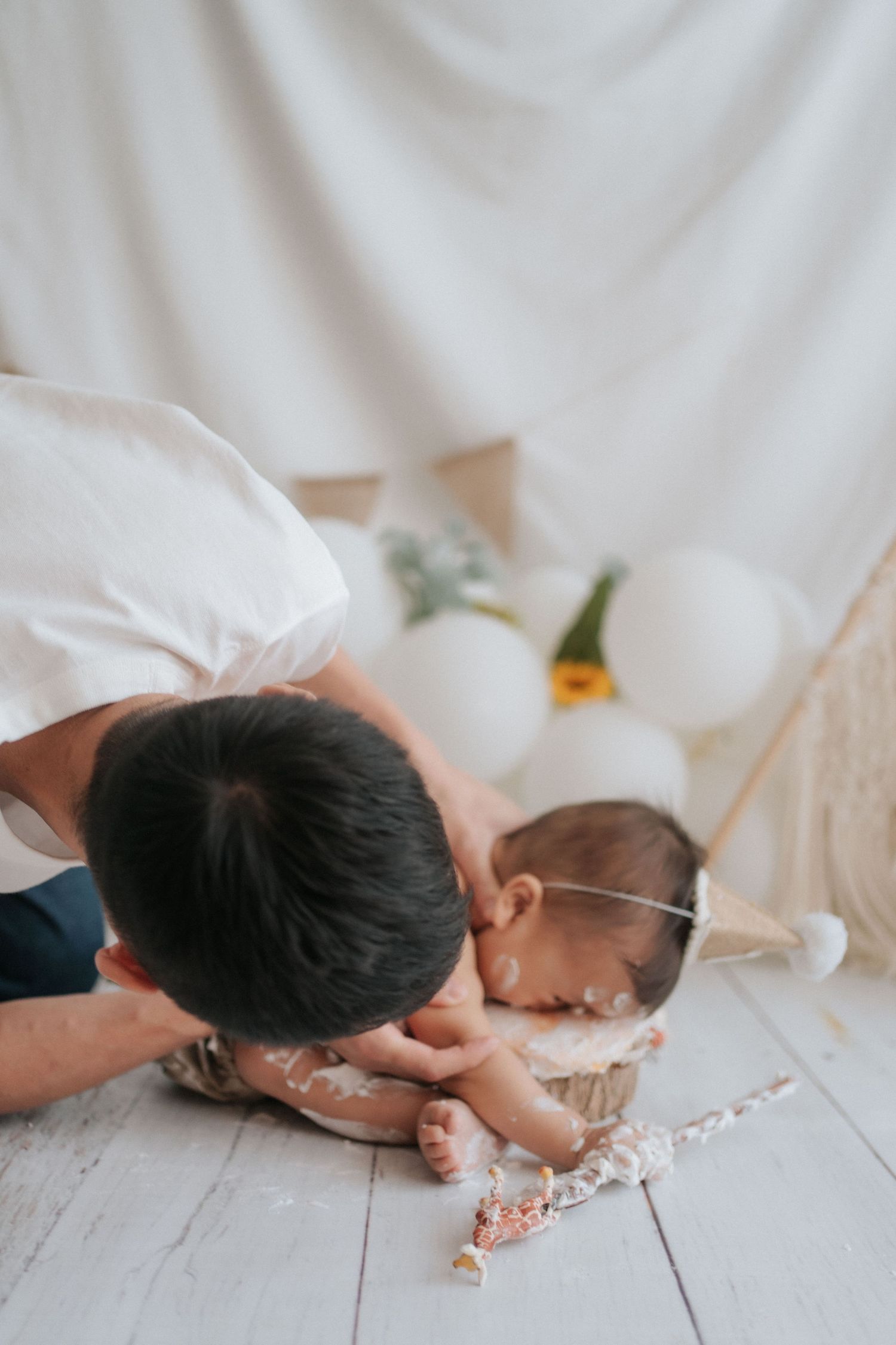A baby lies on a white sheet while playing with pink flower petals scattered on the floor.