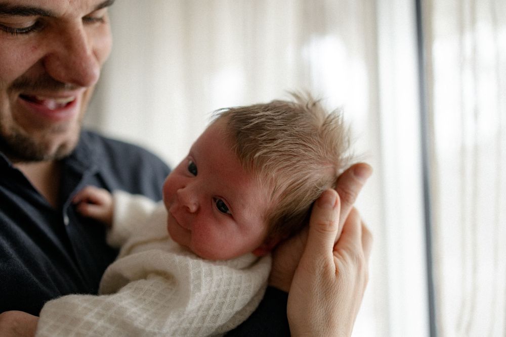 A newborn baby wearing white is gently cradled in someone's hands near a window with soft lighting.