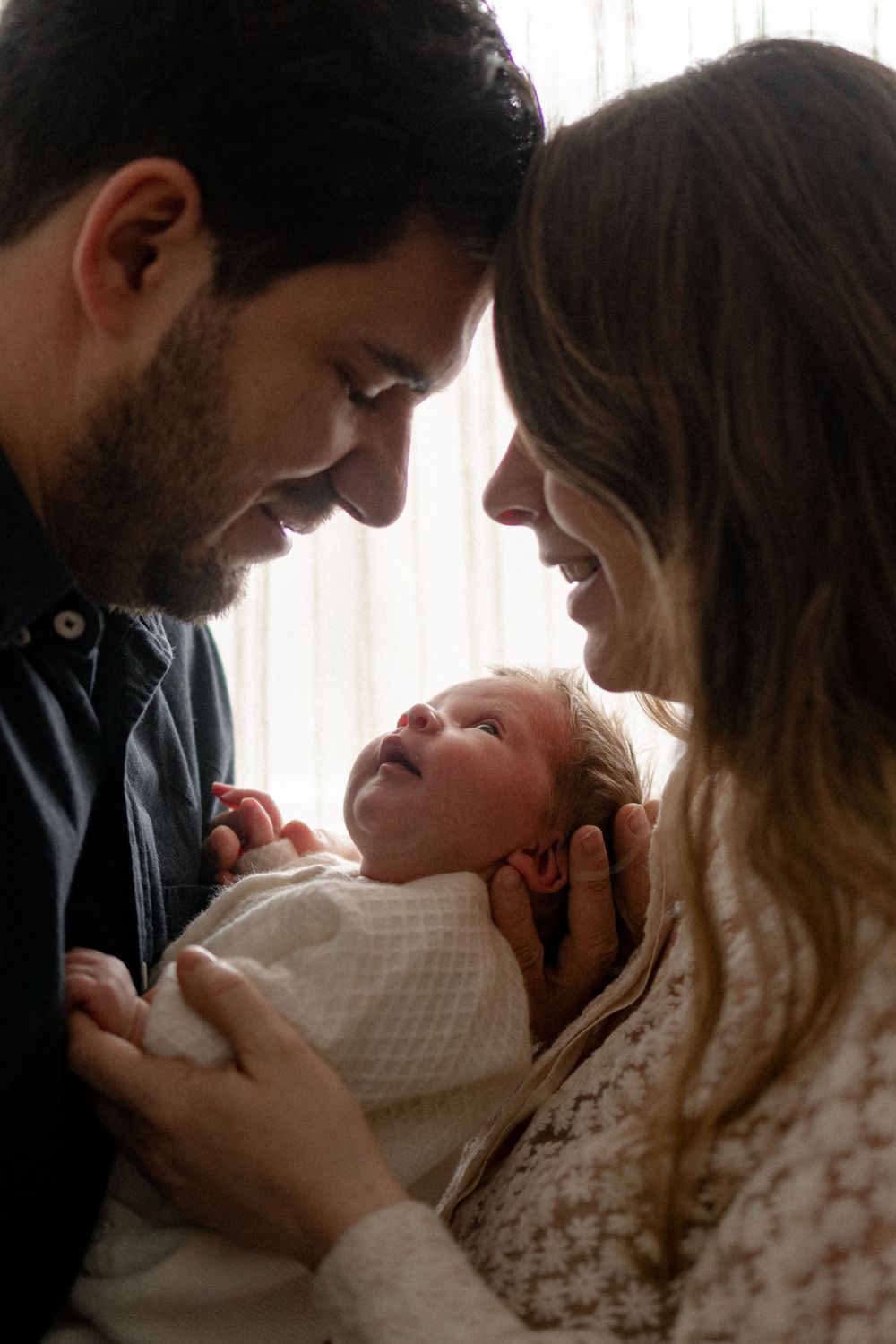 A couple shares tender moments with their newborn baby in a series of intimate photos by a window.