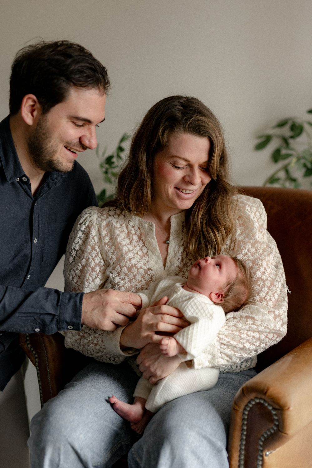 A couple sits together on a brown leather couch, lovingly gazing at their newborn baby wrapped in white.