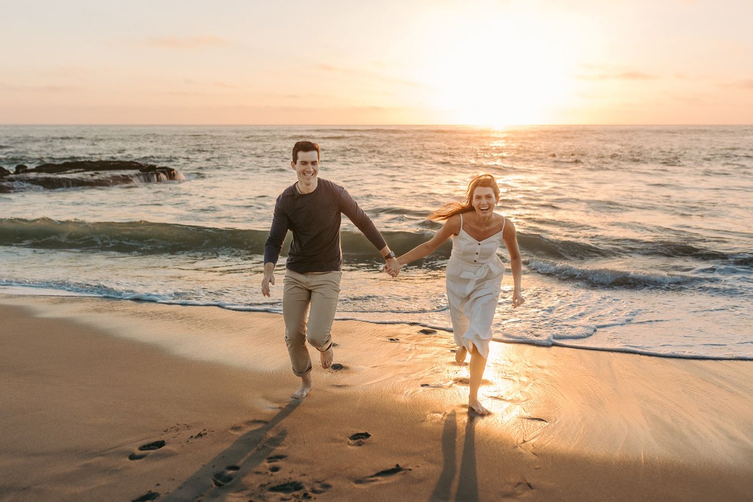 A couple runs hand in hand along Windansea Beach at sunset with crashing waves behind them.