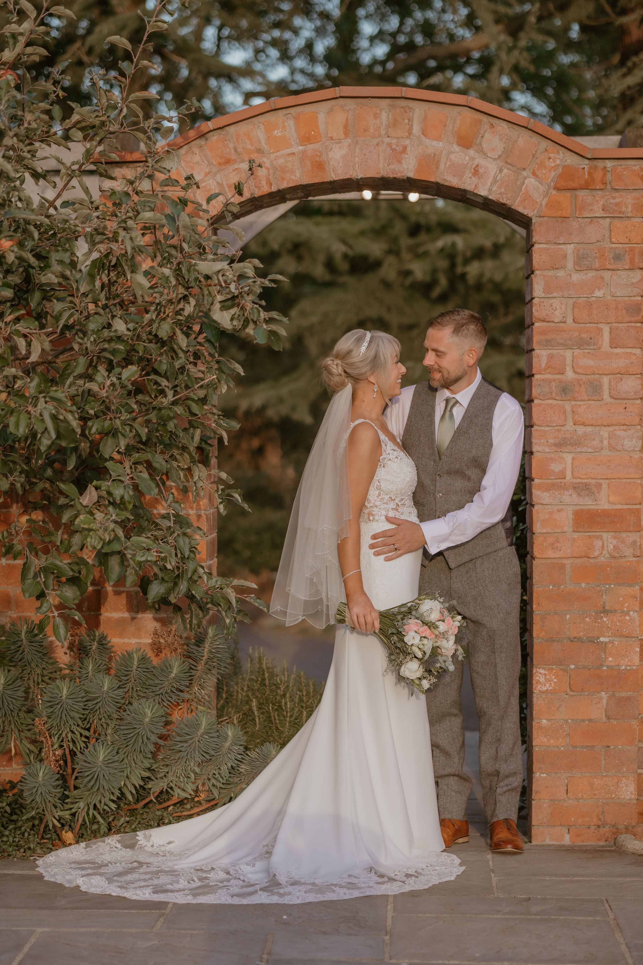 Bride and groom pose under brick archway in garden setting.