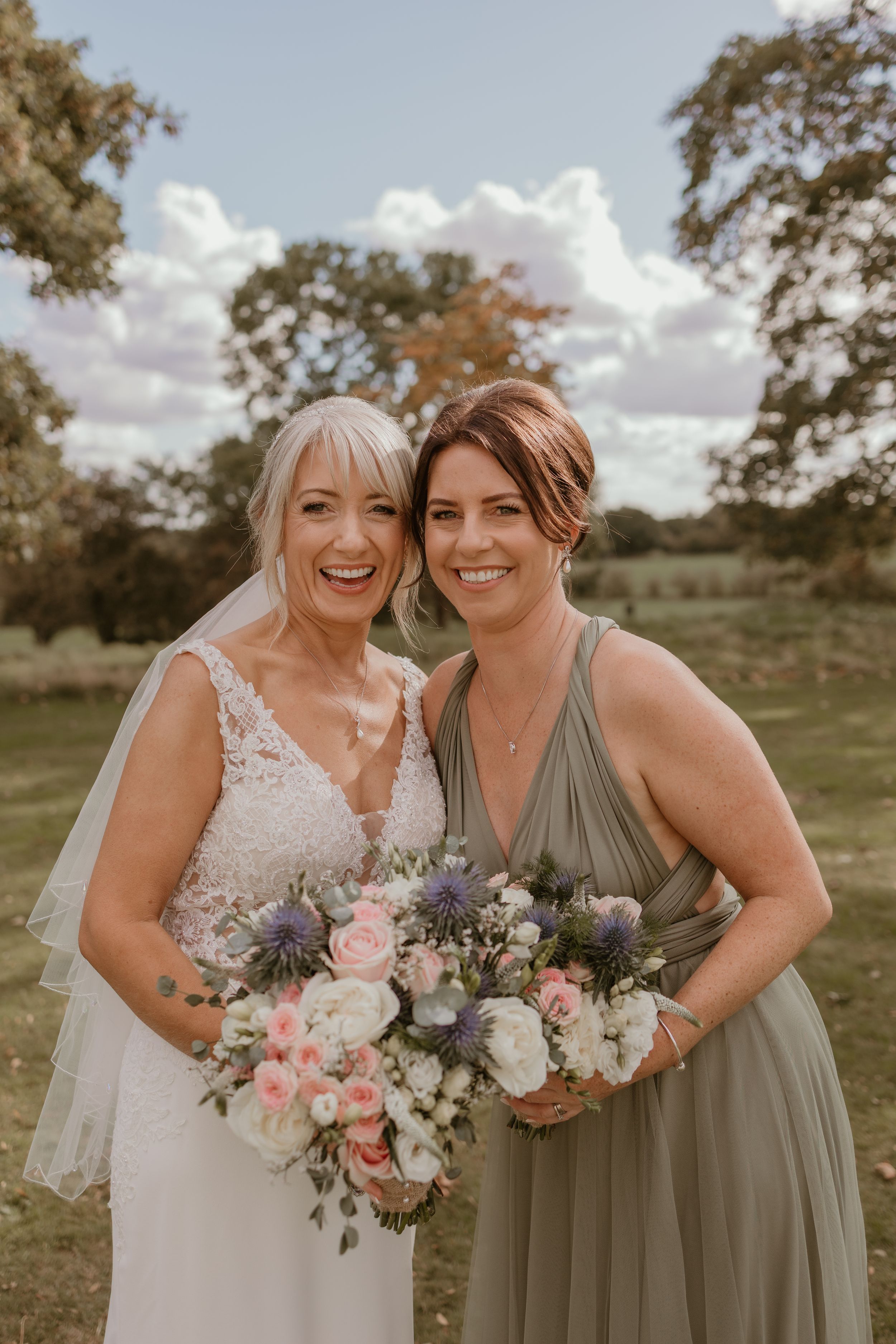 Bride and bridesmaid hold beautiful pastel bouquets while smiling outdoors.