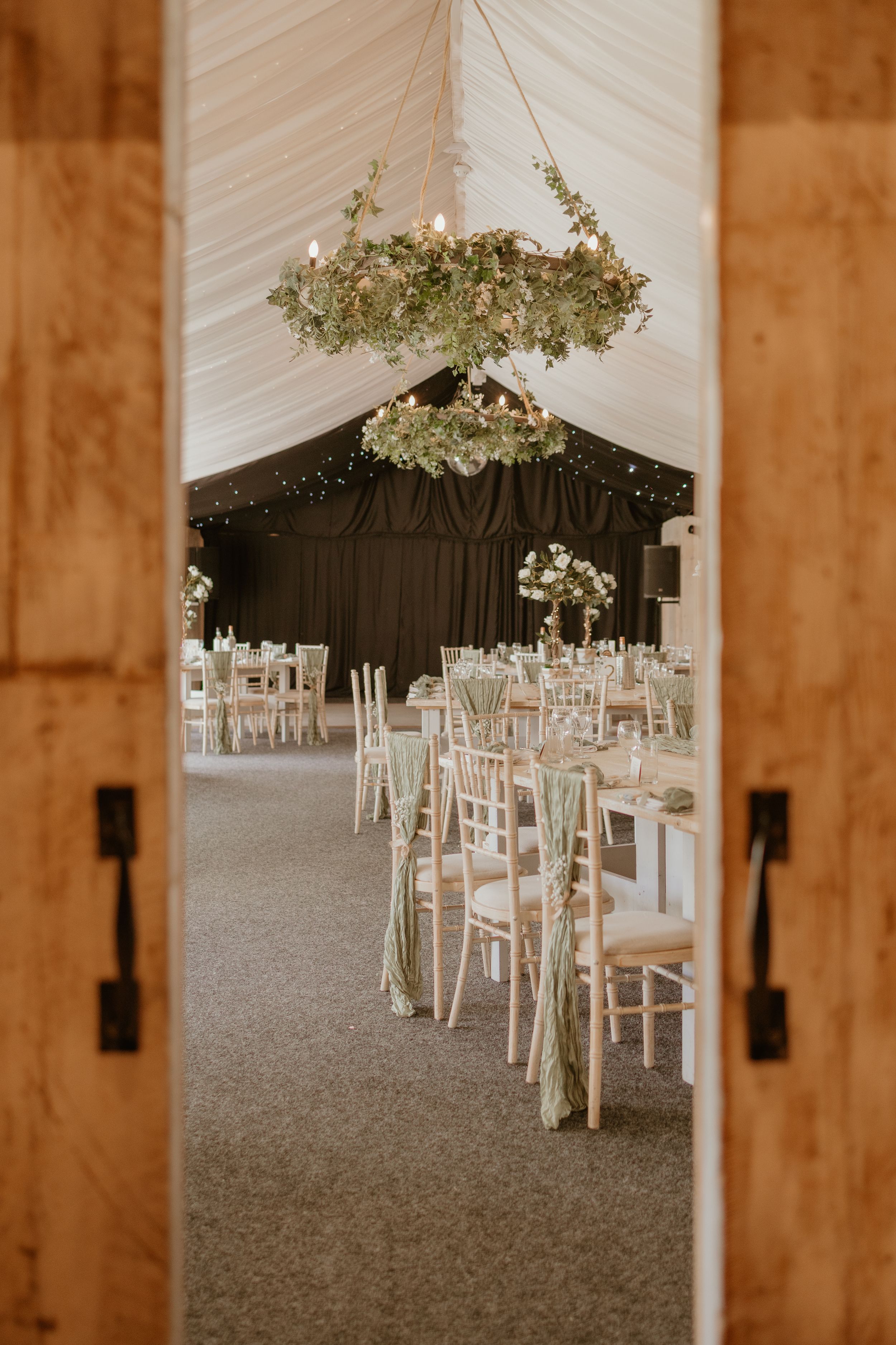Elegant wedding reception setup with floral chandelier and white chairs viewed through wooden double doors. At Ashton Lodge, Rugby.