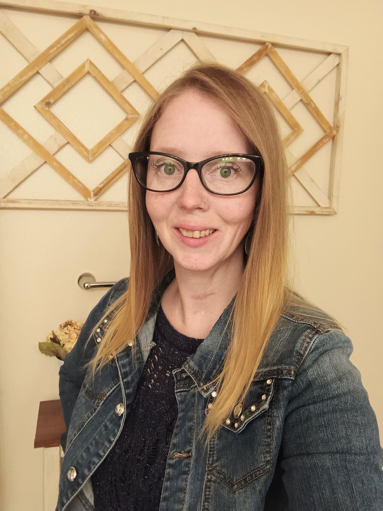 Person wearing glasses and denim jacket poses against geometric wall art.