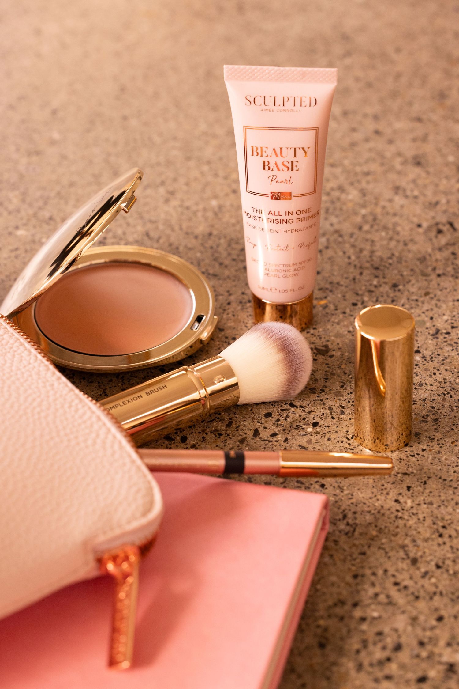 Flatlay of luxury beauty products including compact powder, brush and pink makeup bag on textured surface.
