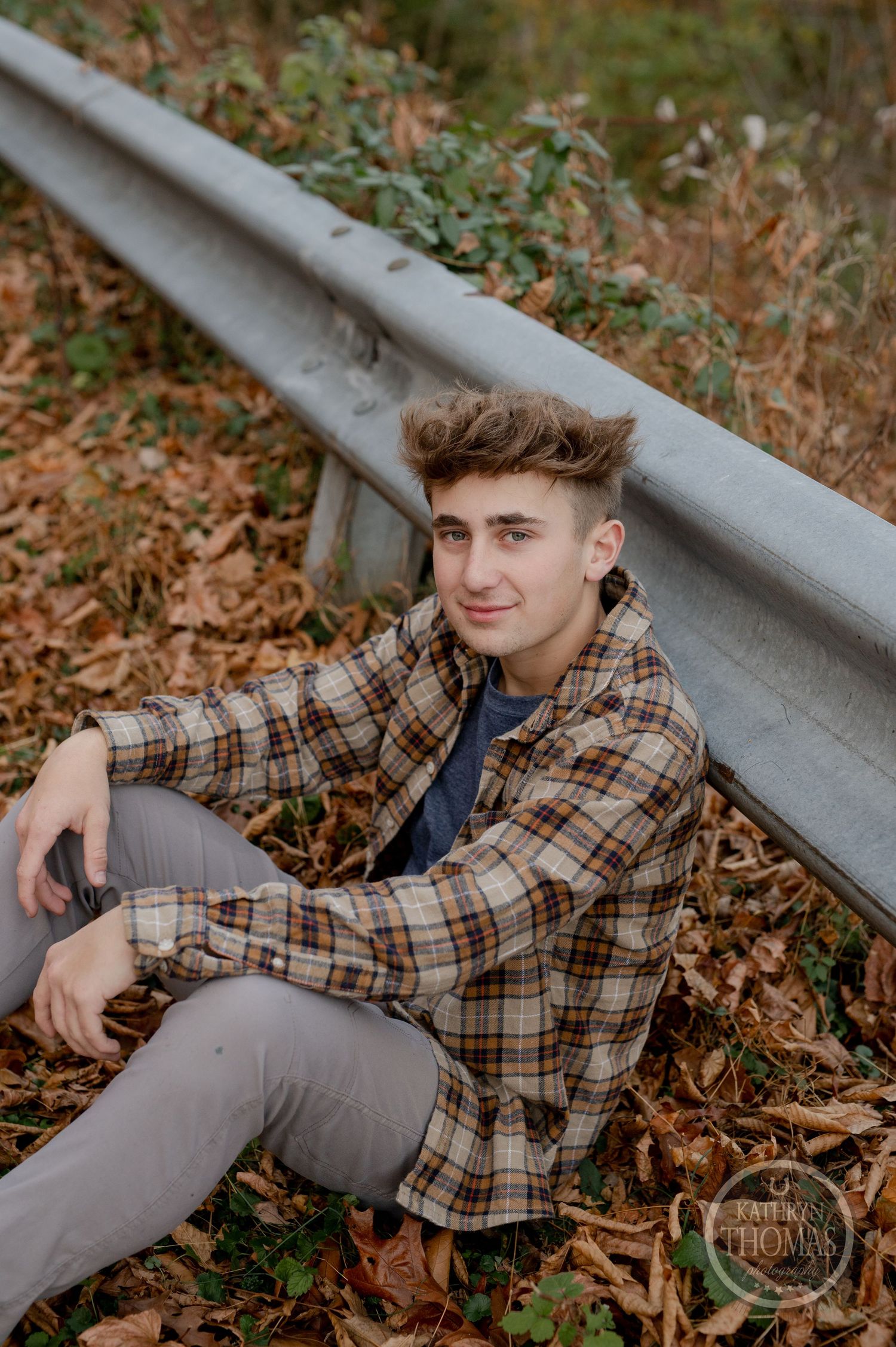 Someone poses casually by a metal guardrail surrounded by fallen autumn leaves and greenery.