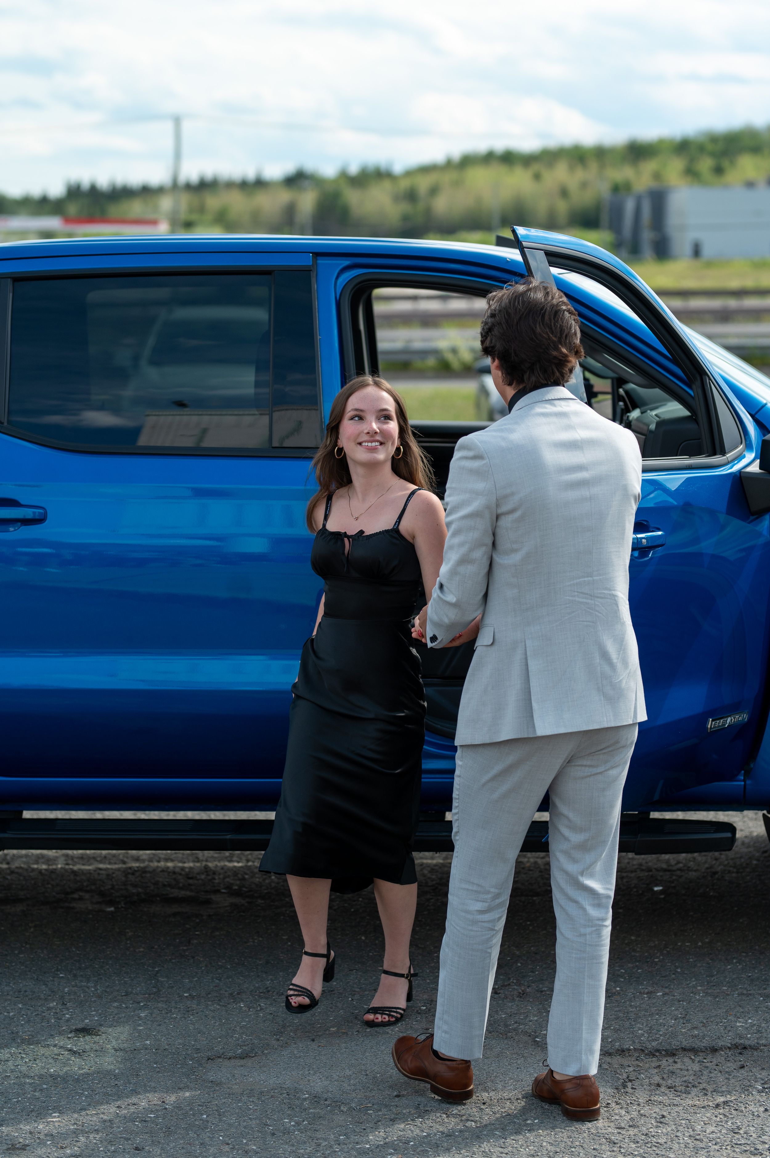 Person in black dress stands by blue vehicle while talking to someone in light suit.