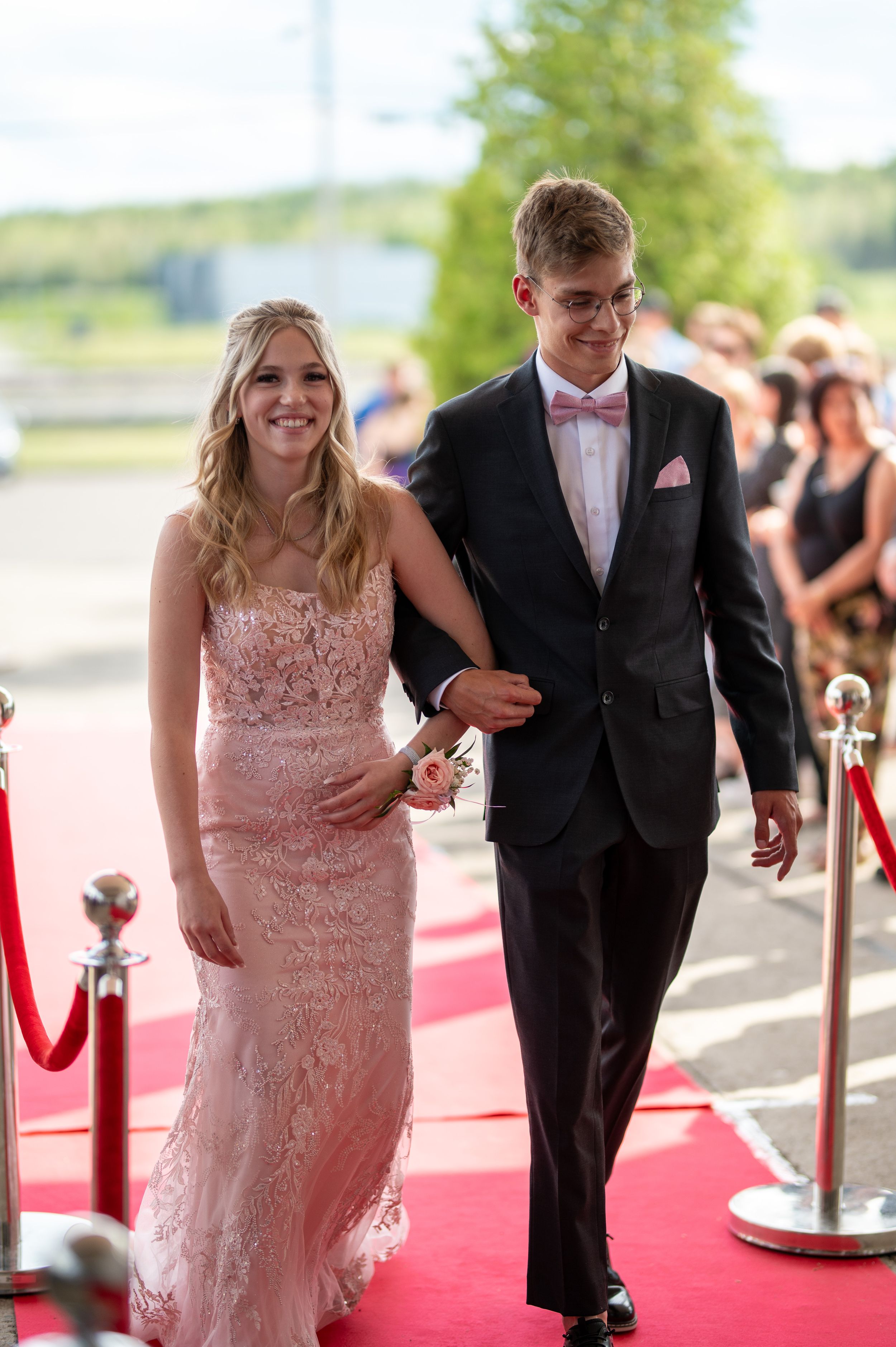 A couple walks down a red carpet at a formal event, wearing a blush pink lace gown and black tuxedo.