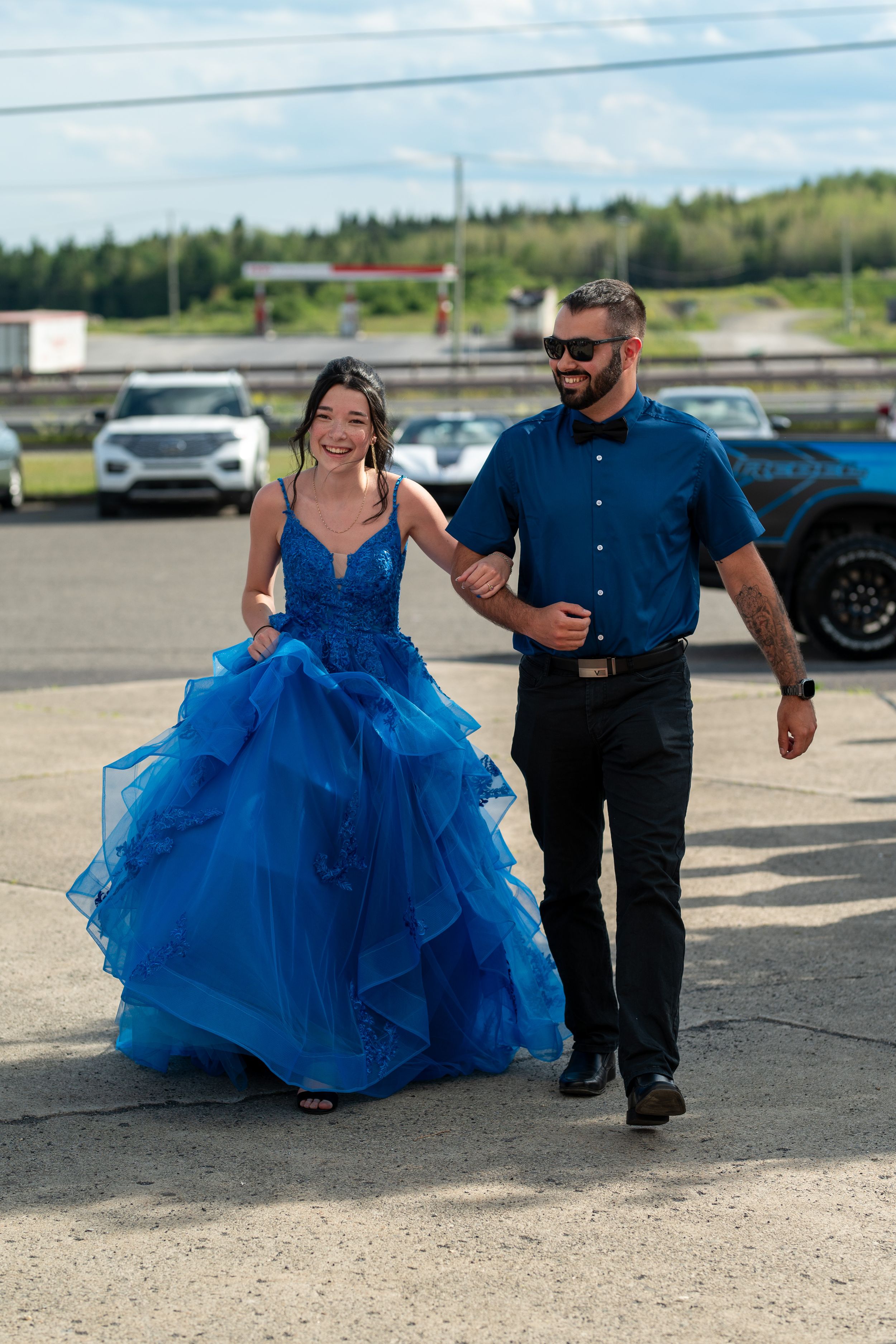 A couple walks through a parking lot, with the woman wearing a flowing royal blue ball gown.