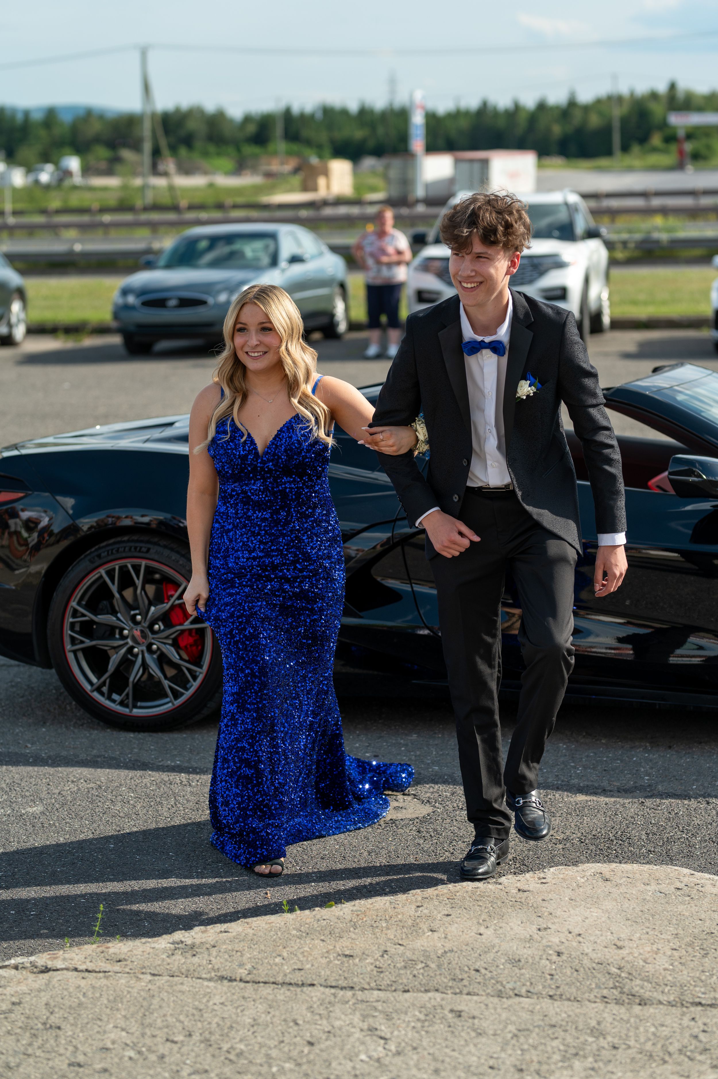 A couple in formal attire walks past luxury cars in a parking lot.