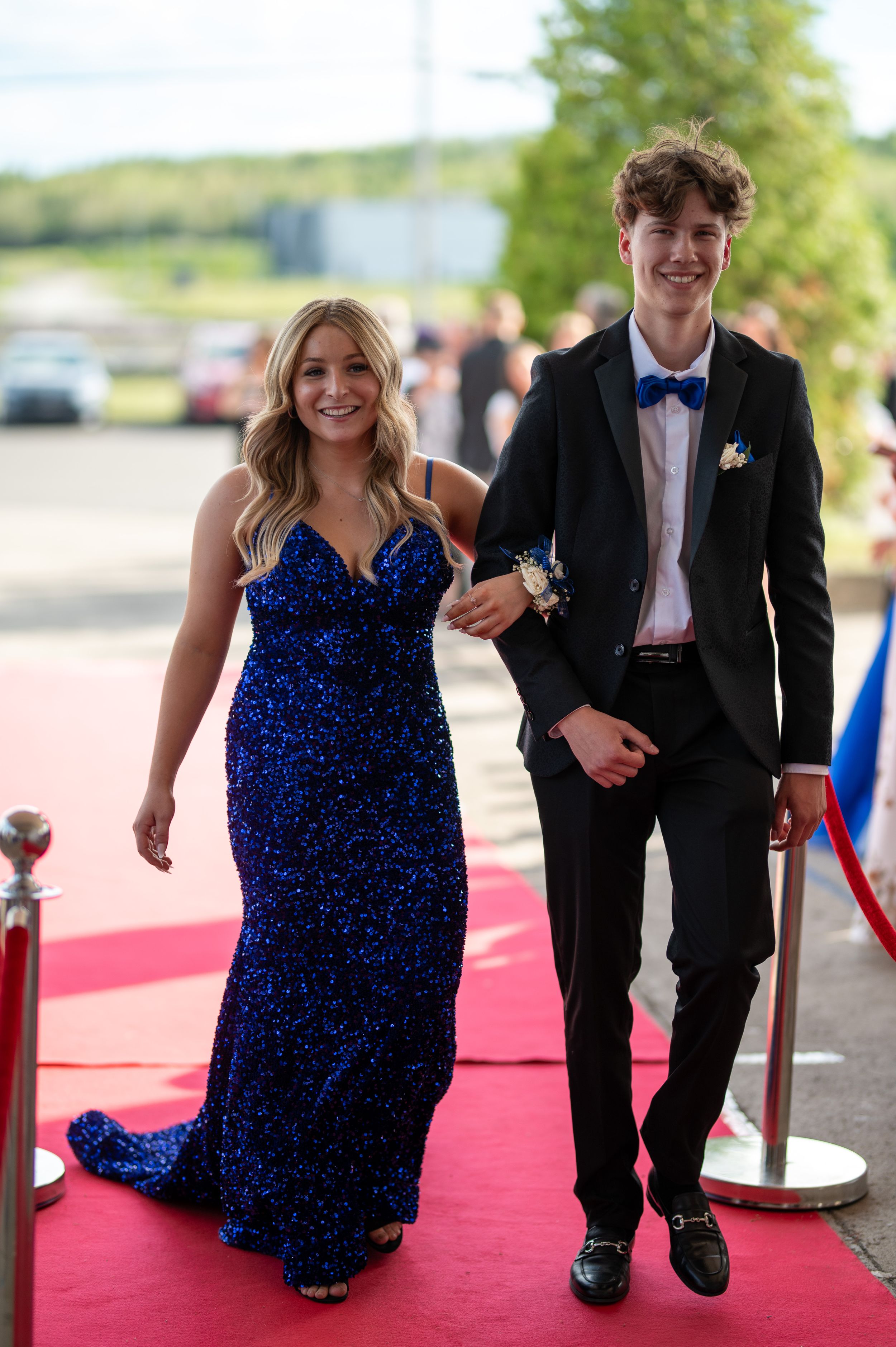 A couple walks arm in arm down a red carpet in matching blue and black formal wear.