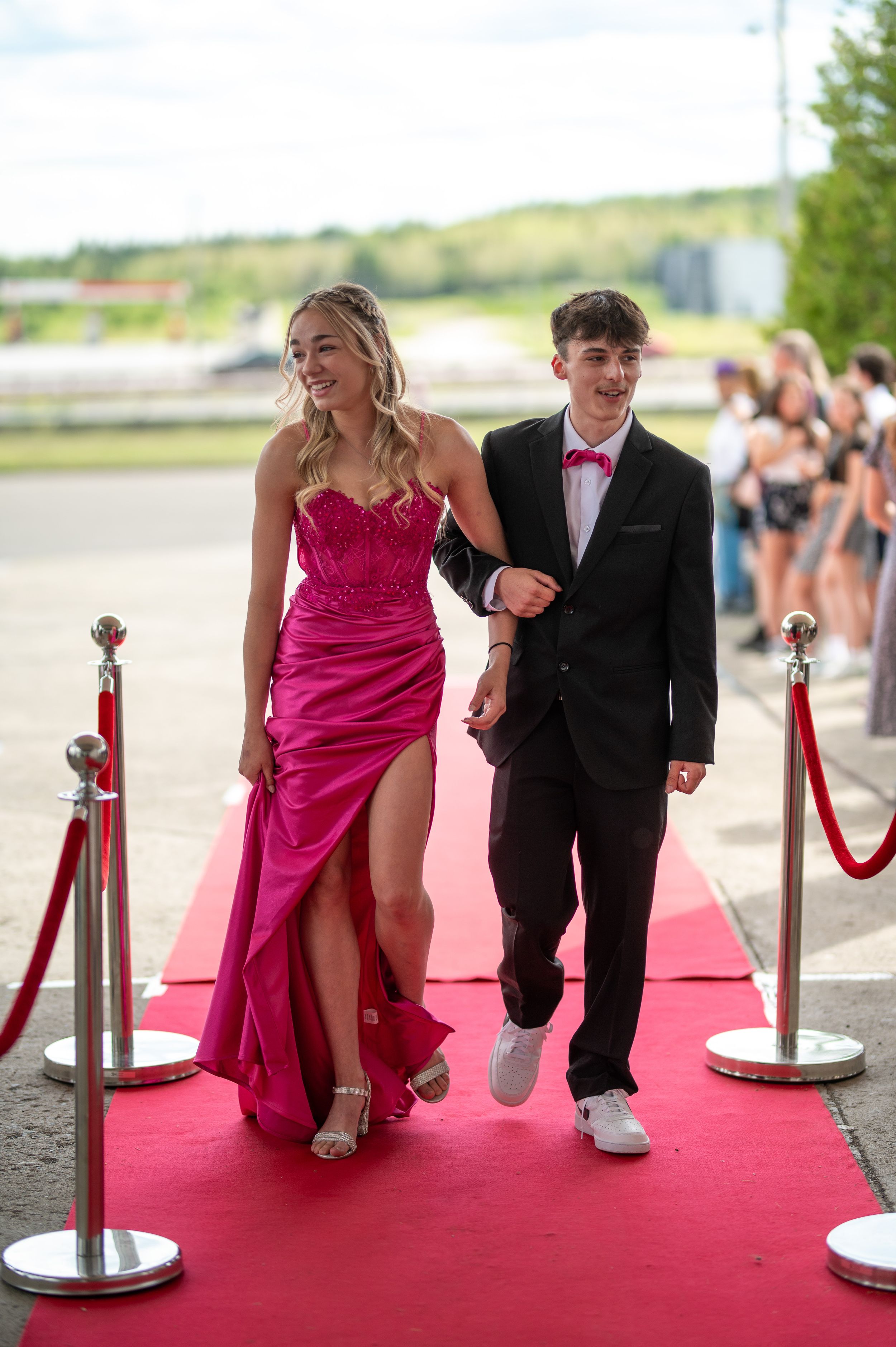 A couple walking on a red carpet, wearing a hot pink gown and black tuxedo with bow tie.