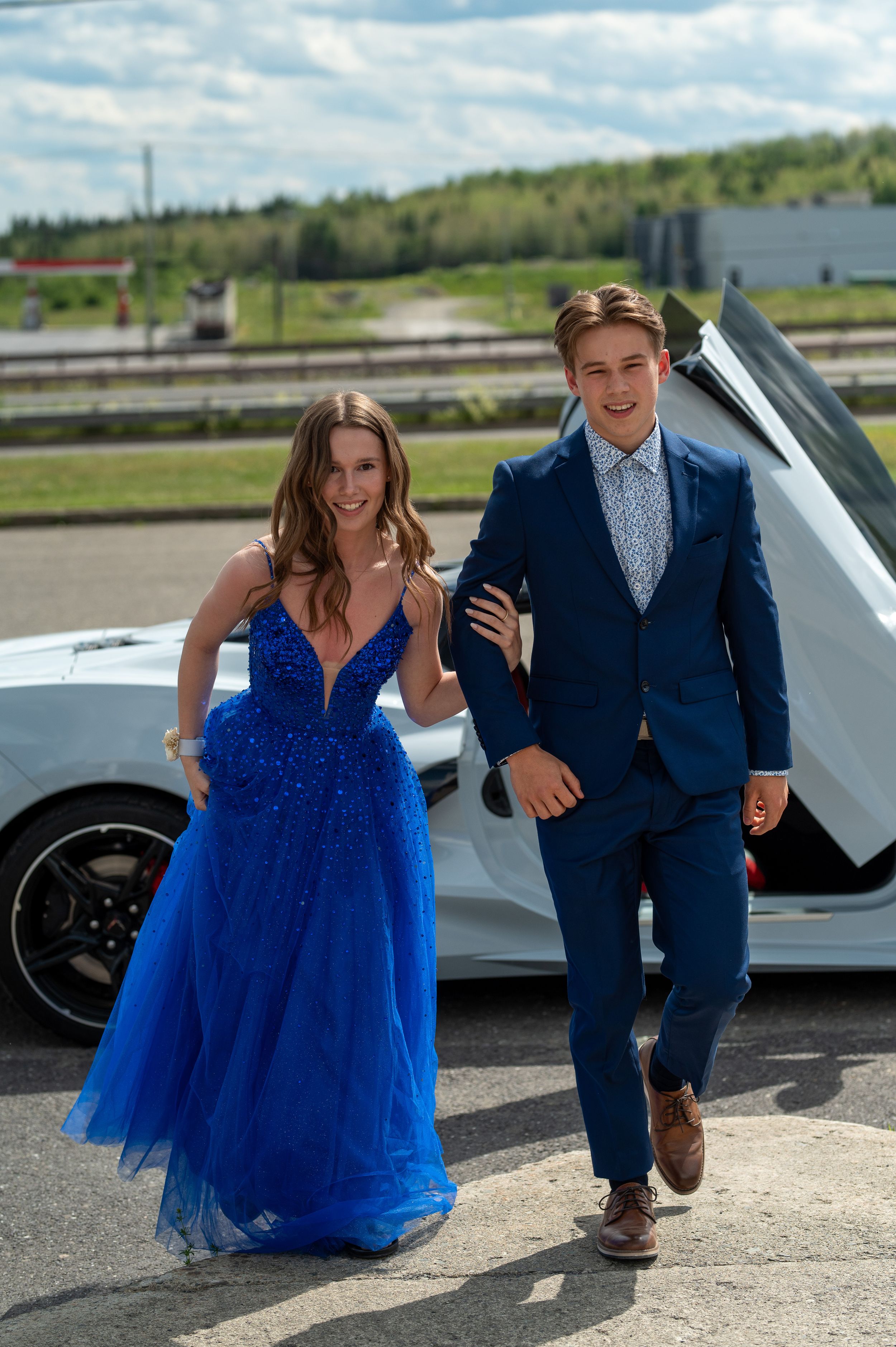 A couple in formal wear - blue gown and navy suit - posing next to a luxury white sports car.