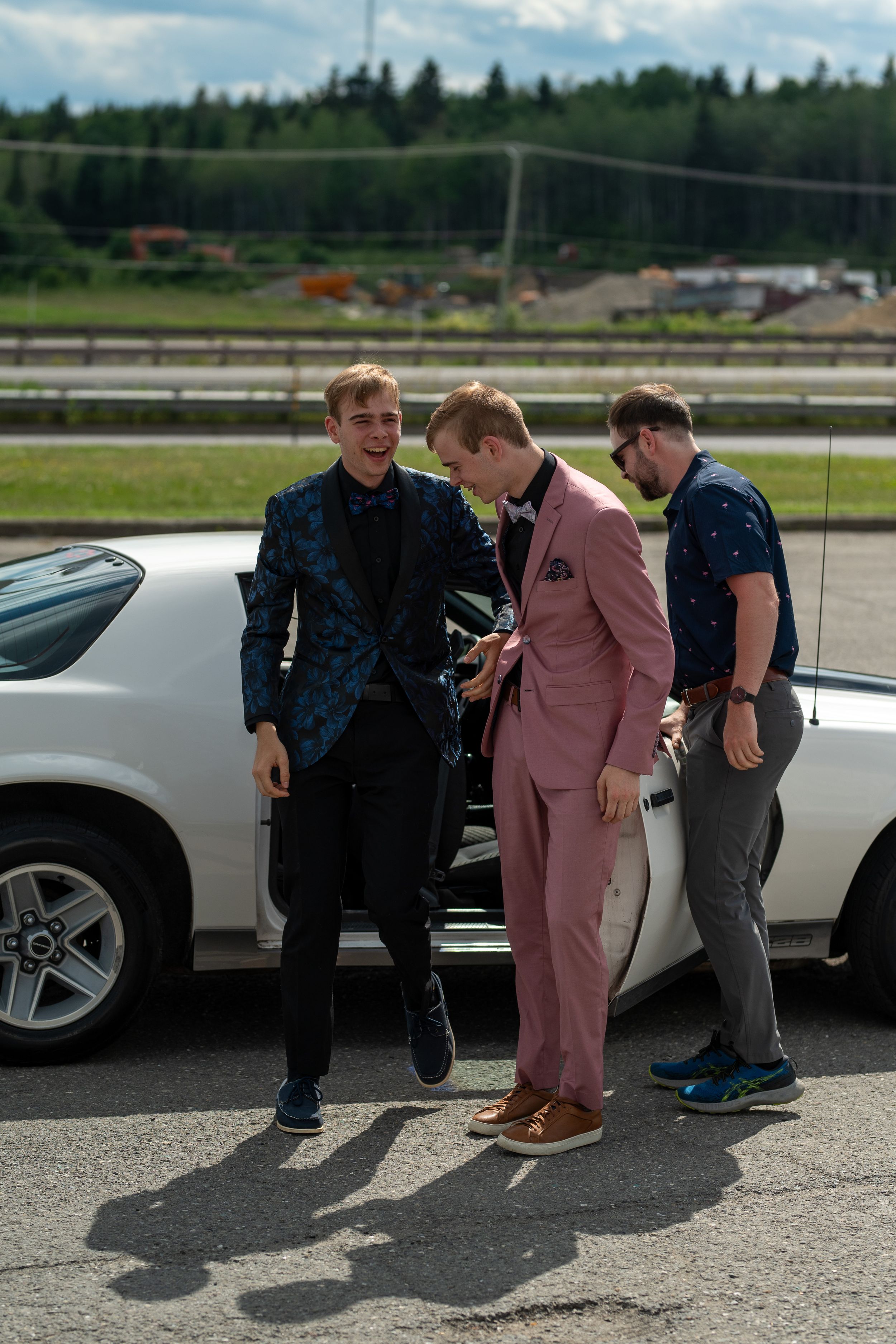 Three young men in formal suits standing by luxury vehicles in a parking lot.