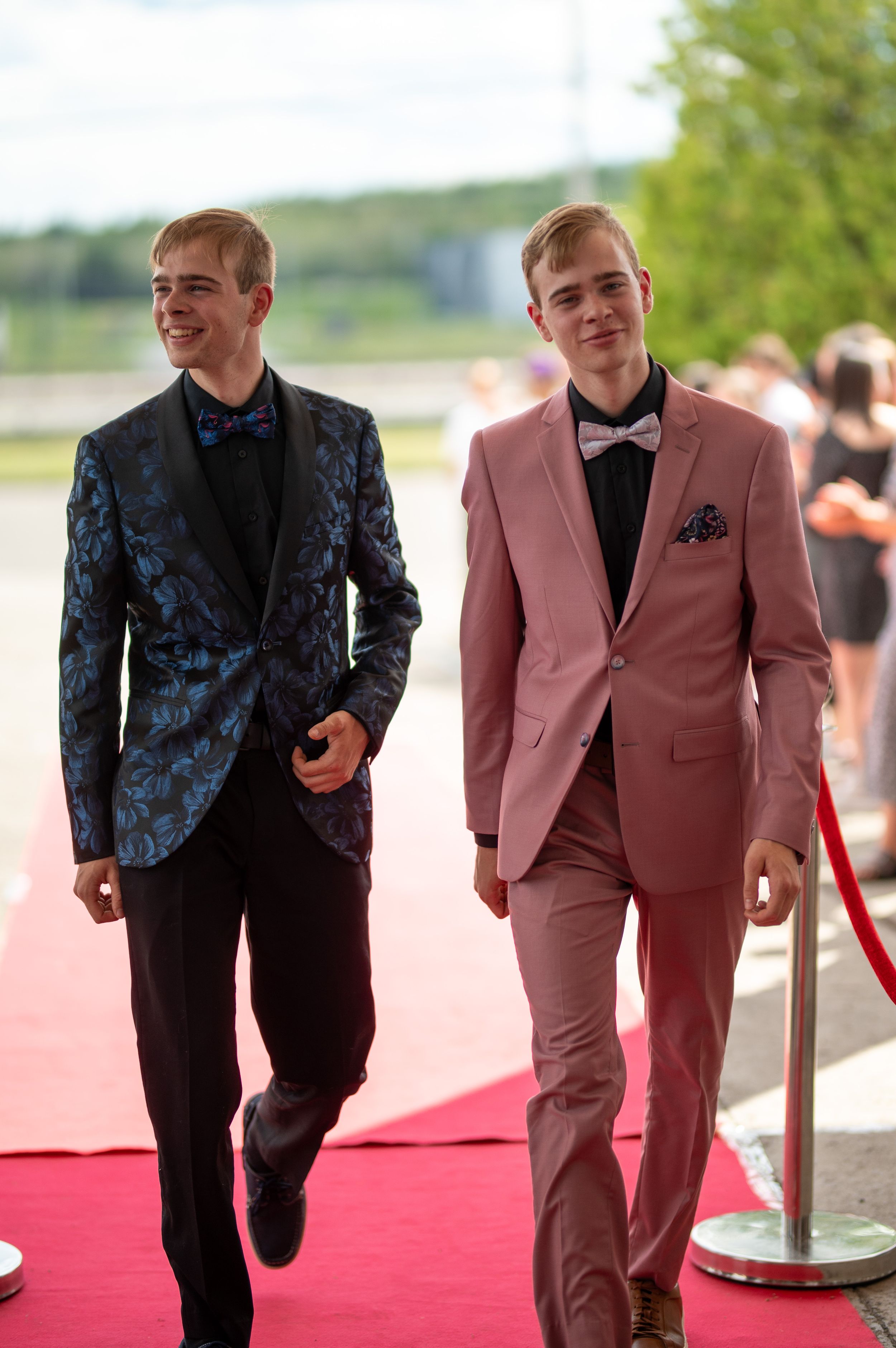Two young men in formal attire walk down a red carpet, one wearing a floral blue blazer and one in pink.