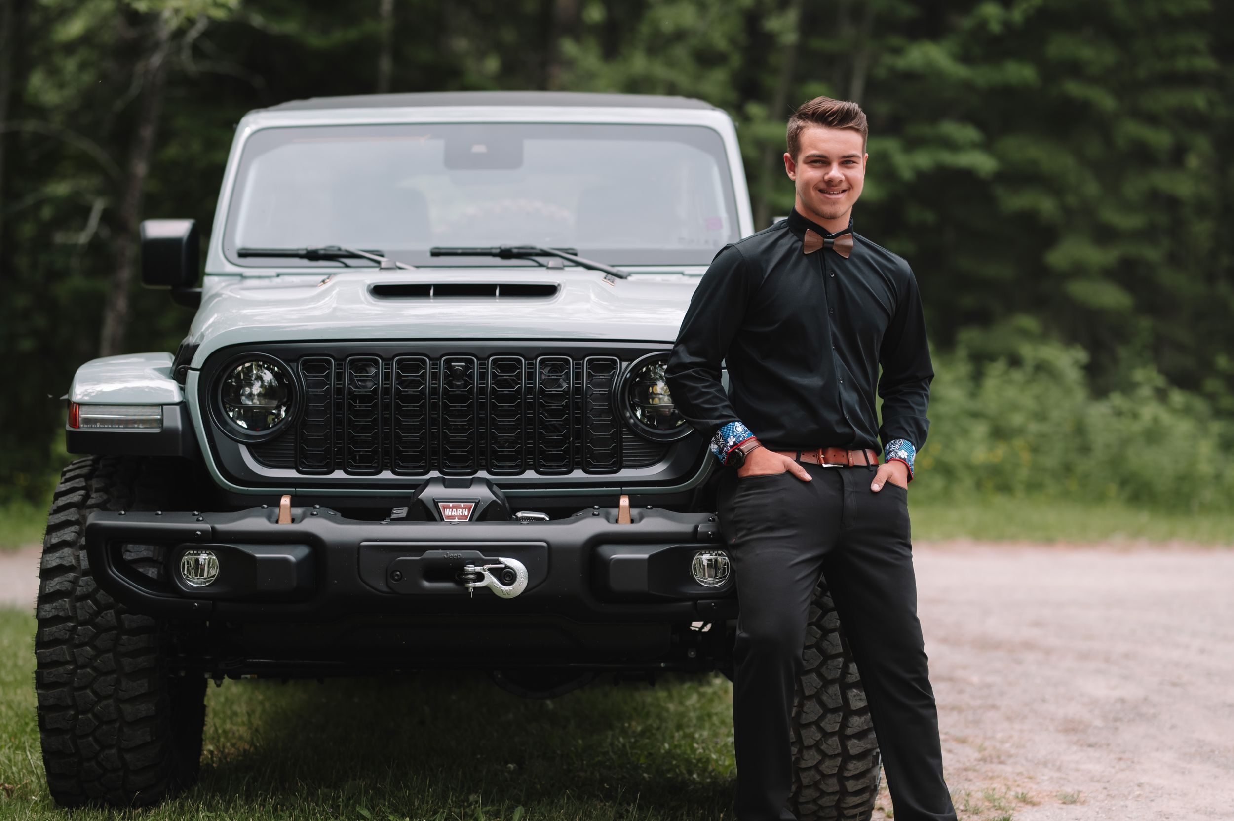 Man in dark shirt poses confidently next to black Jeep Wrangler on forest road.