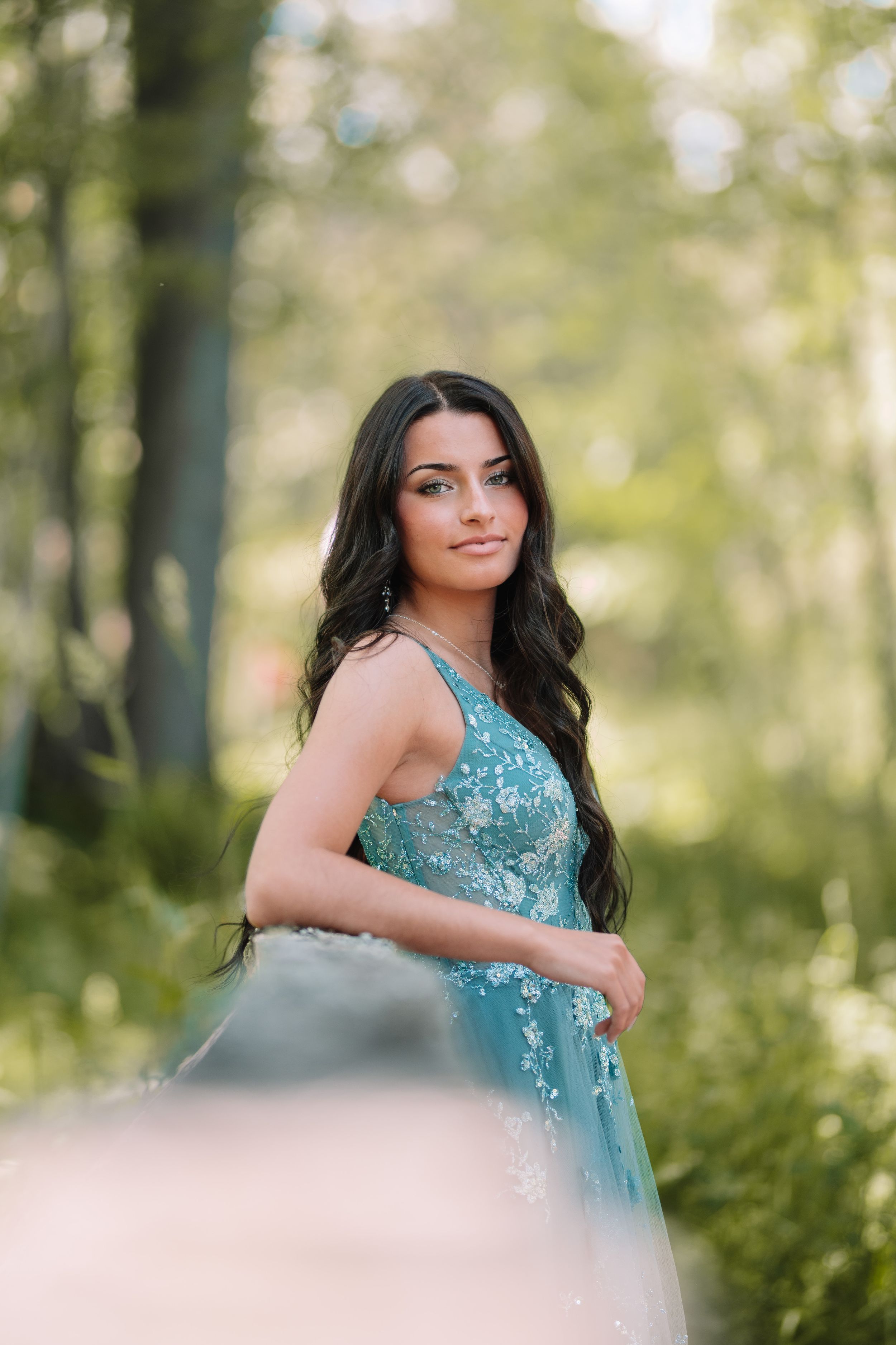 Woman in aqua formal dress poses gracefully in sunlit wooded setting.