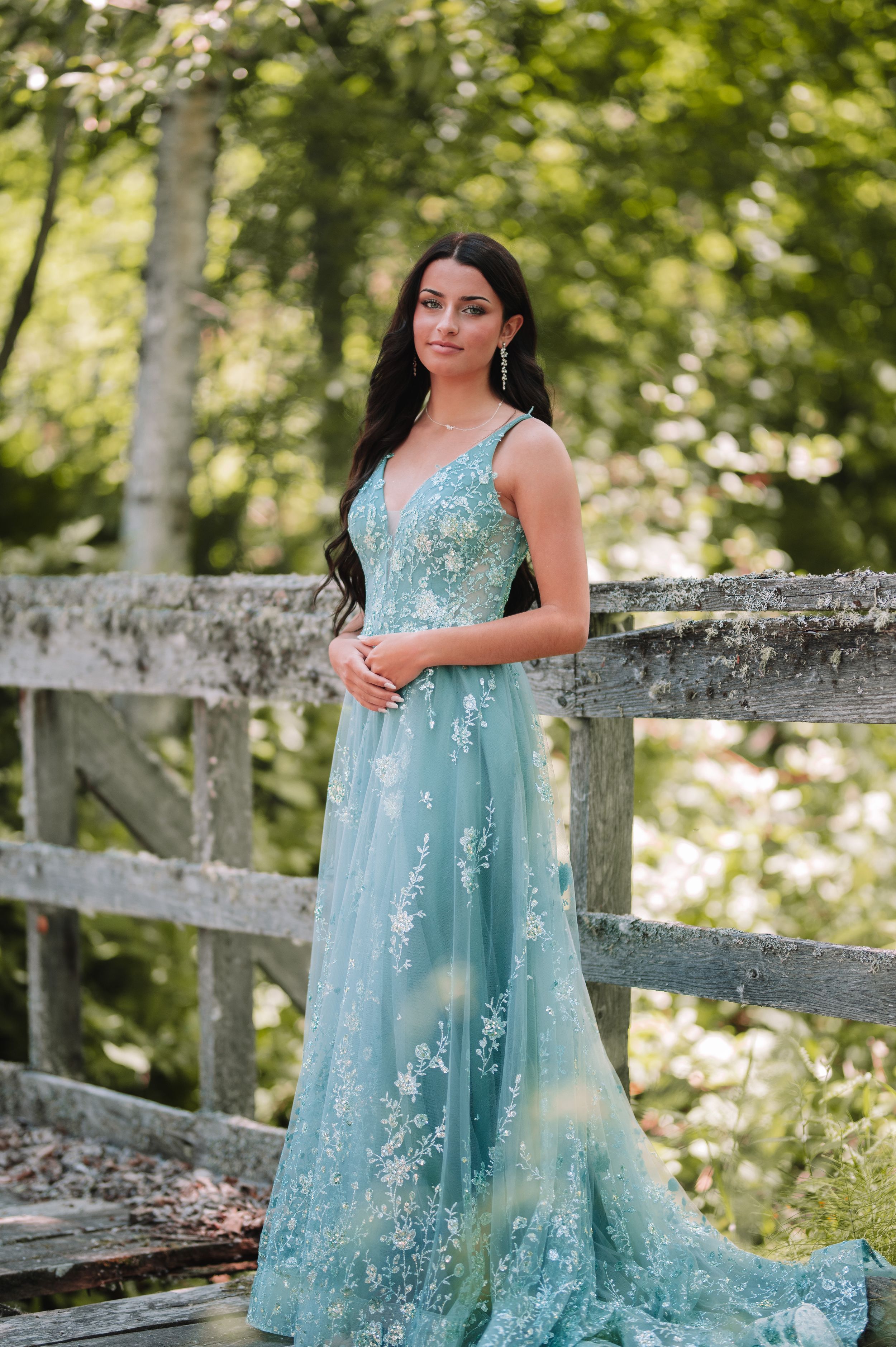 A turquoise blue formal gown with floral embellishments photographed on a wooden bridge in a forest setting.