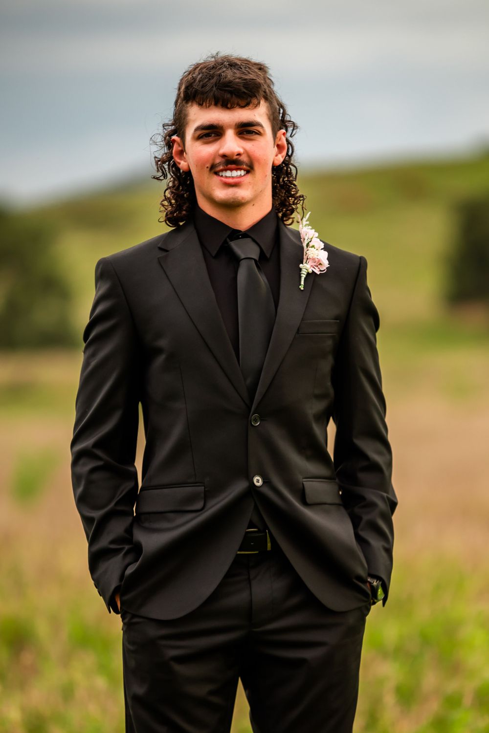 Person wearing black formal suit with boutonniere in a grassy field.