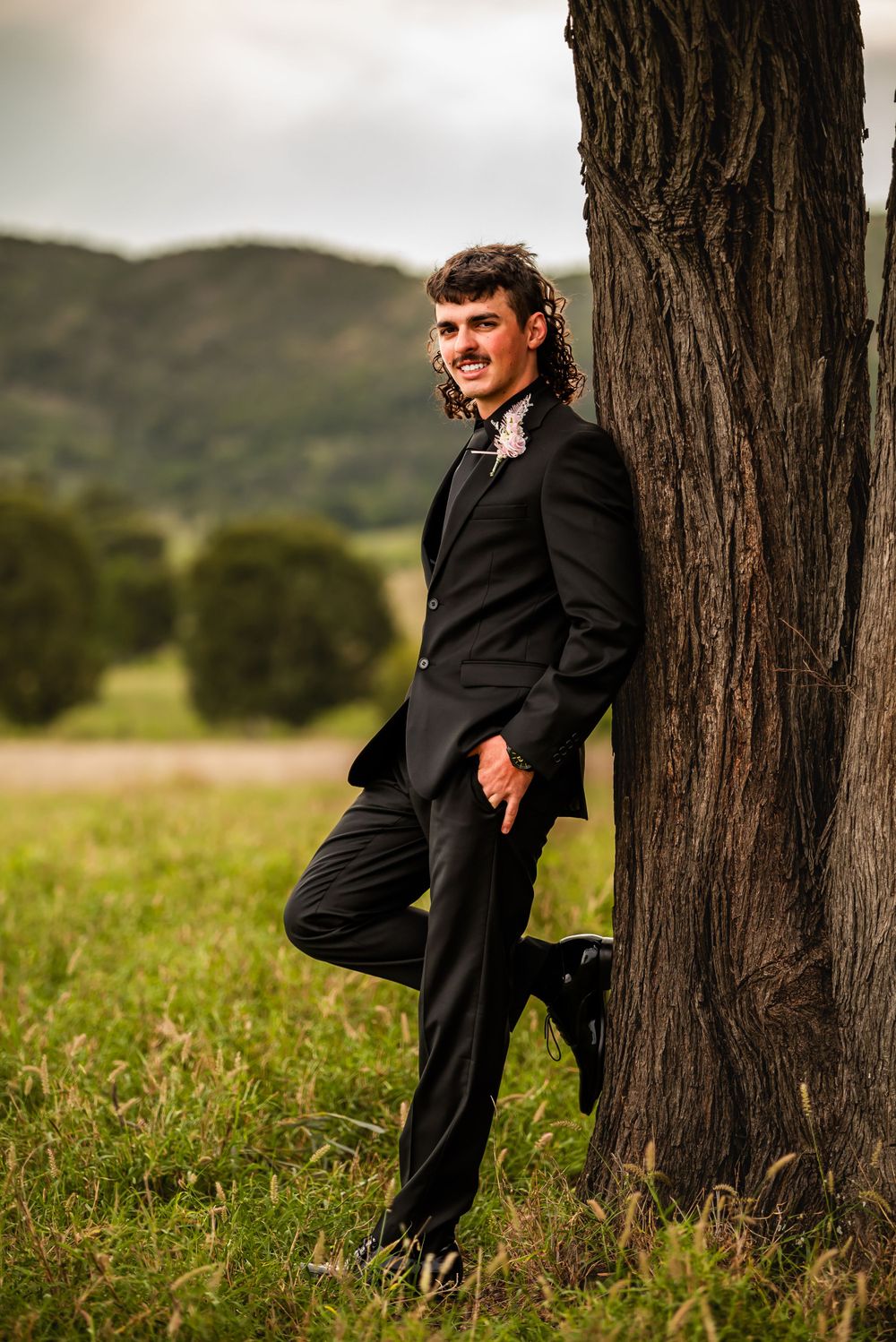 Person in formal black suit leaning against large tree trunk in countryside.
