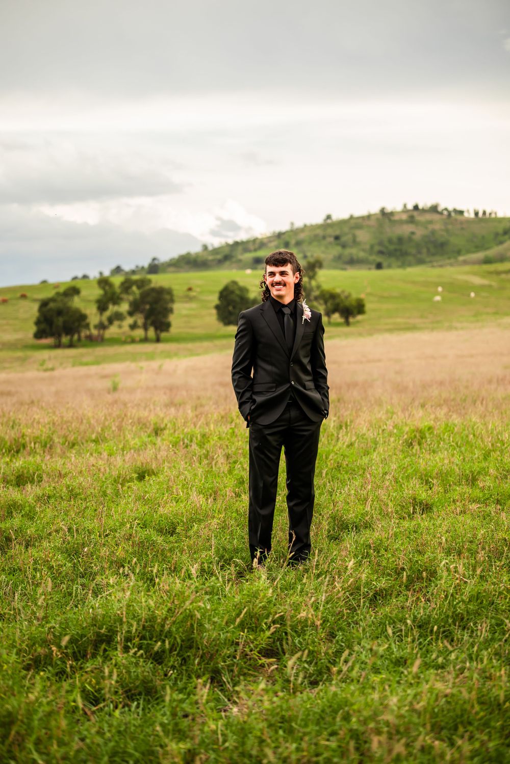 Person in black suit standing in grassy field with rolling hills in background.