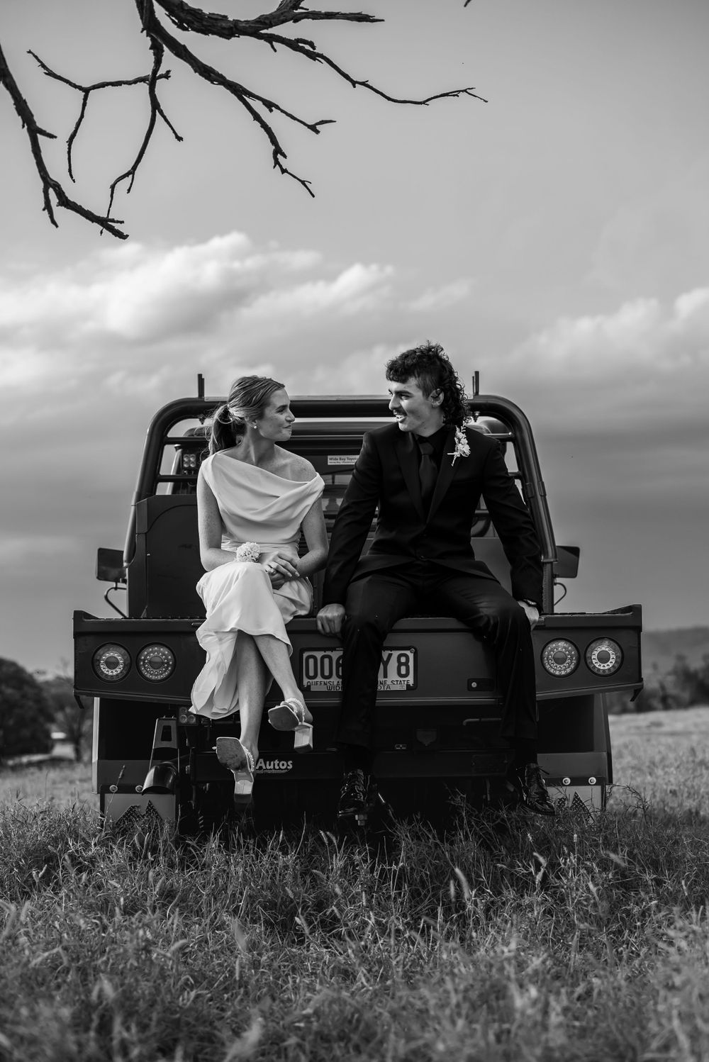 A couple sits together on the back of a vintage truck in a scenic outdoor setting with dramatic clouds overhead.