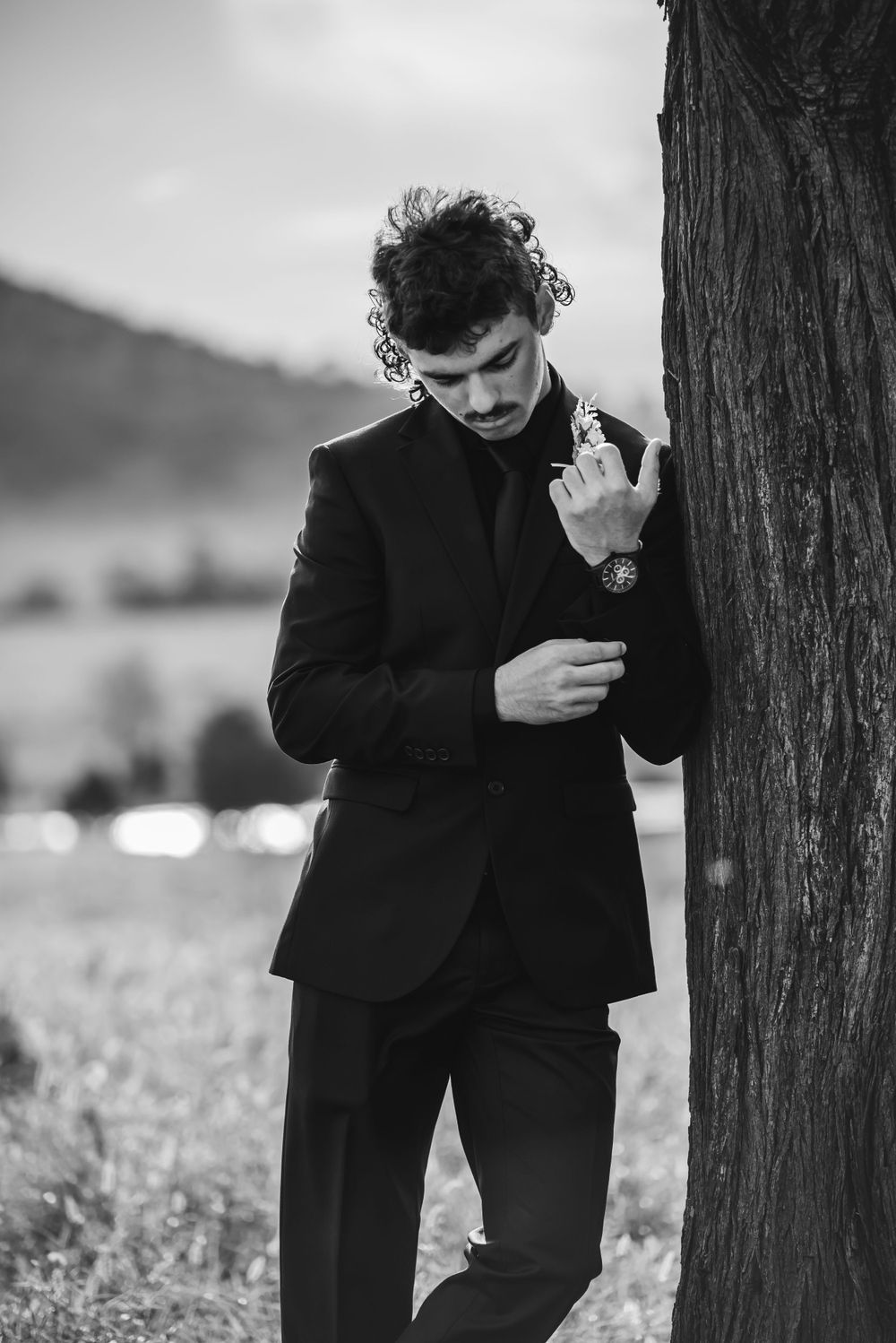 A person in a black suit leaning against a tree while adjusting their cufflinks.