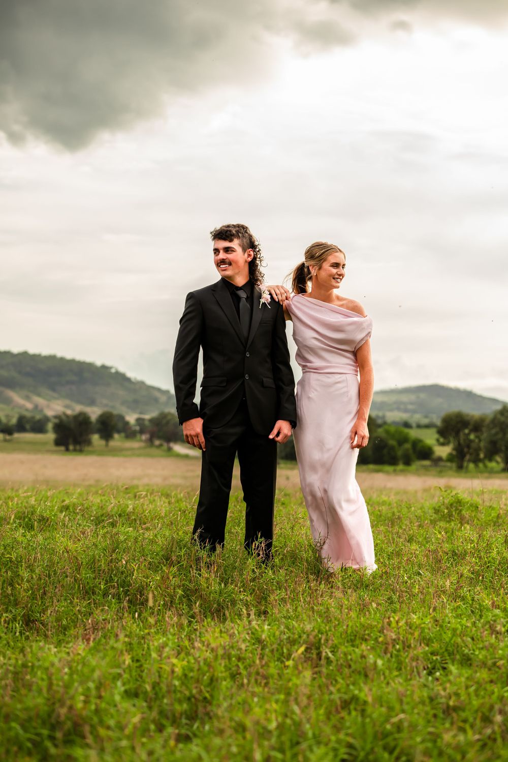 A couple in formal attire standing together in a scenic meadow with rolling hills behind them.