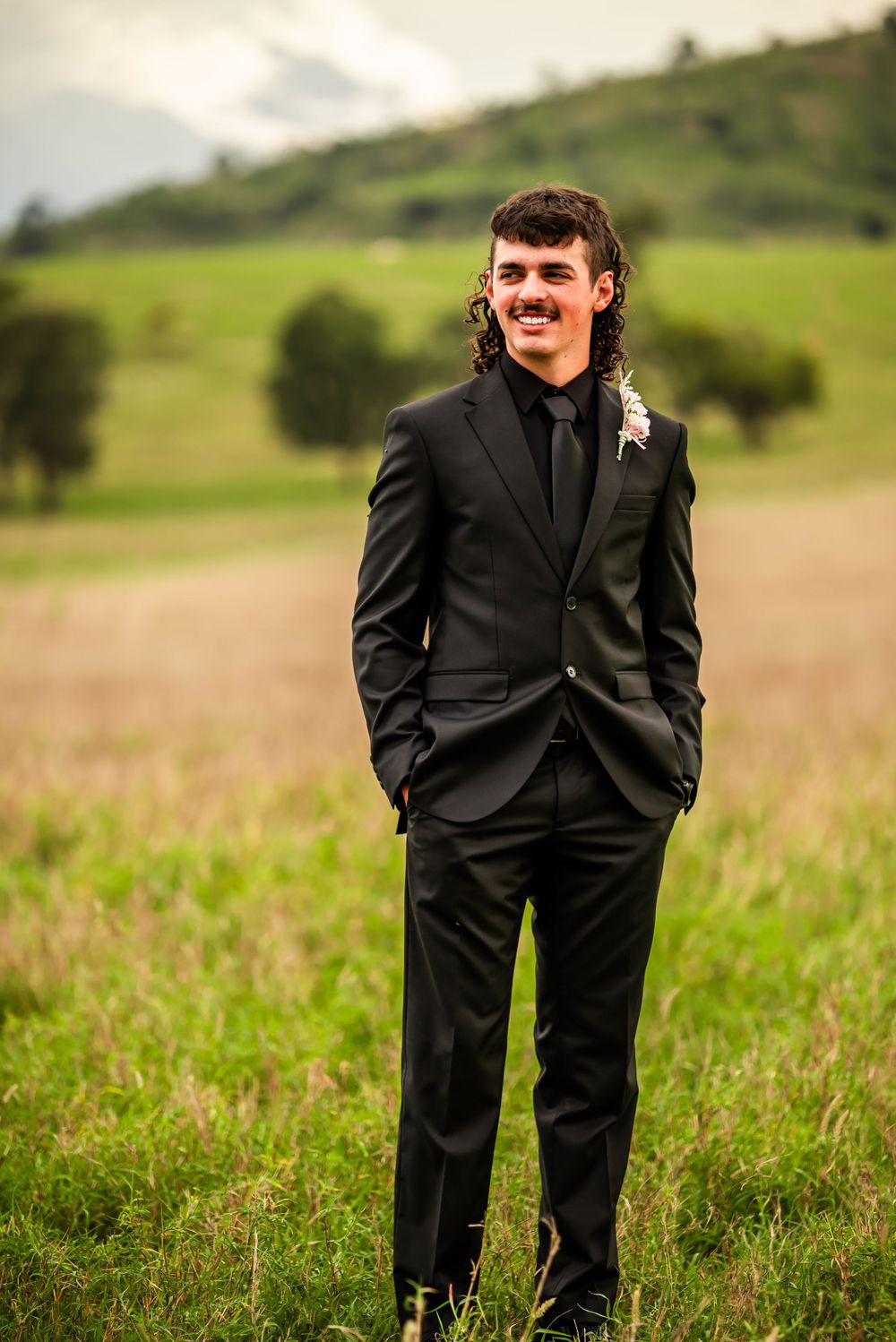 A person in a black suit smiling while standing in a grassy field with hills in the background.