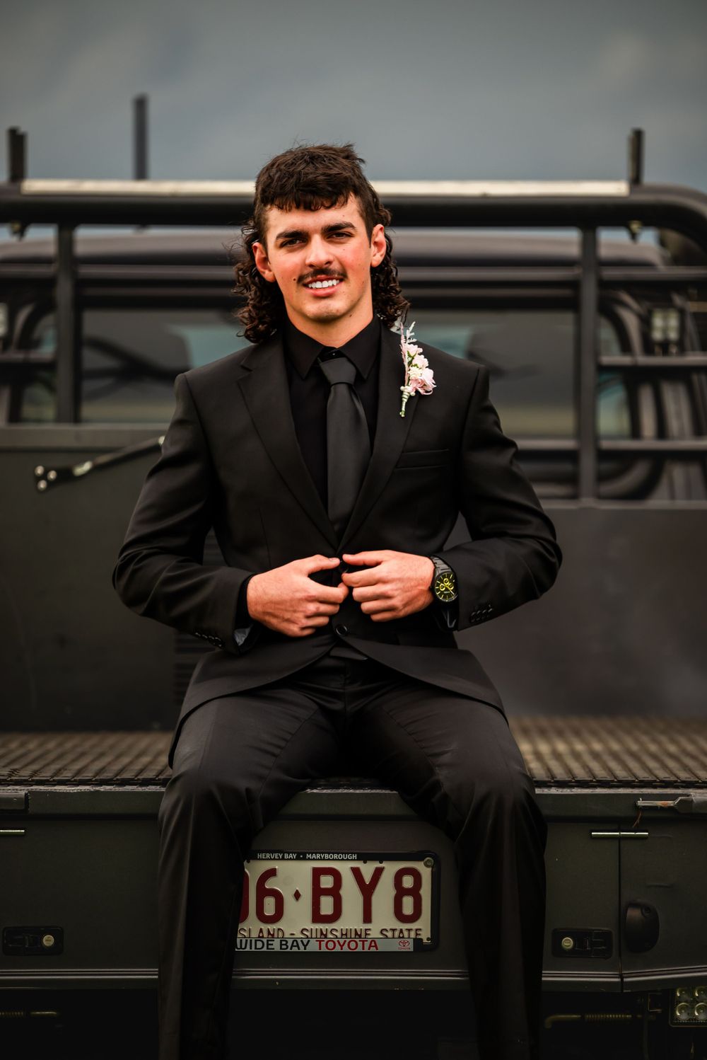 A person in formal attire sitting on the bed of a vintage truck with a visible license plate.