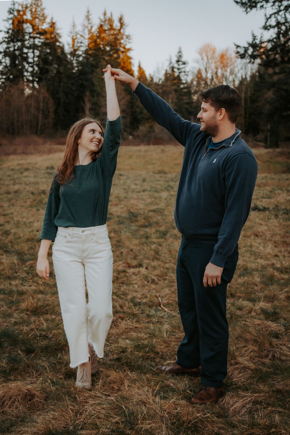 A man twirls a smiling woman in white pants by the hand in a grassy field with evergreen trees at an engagement photo session.