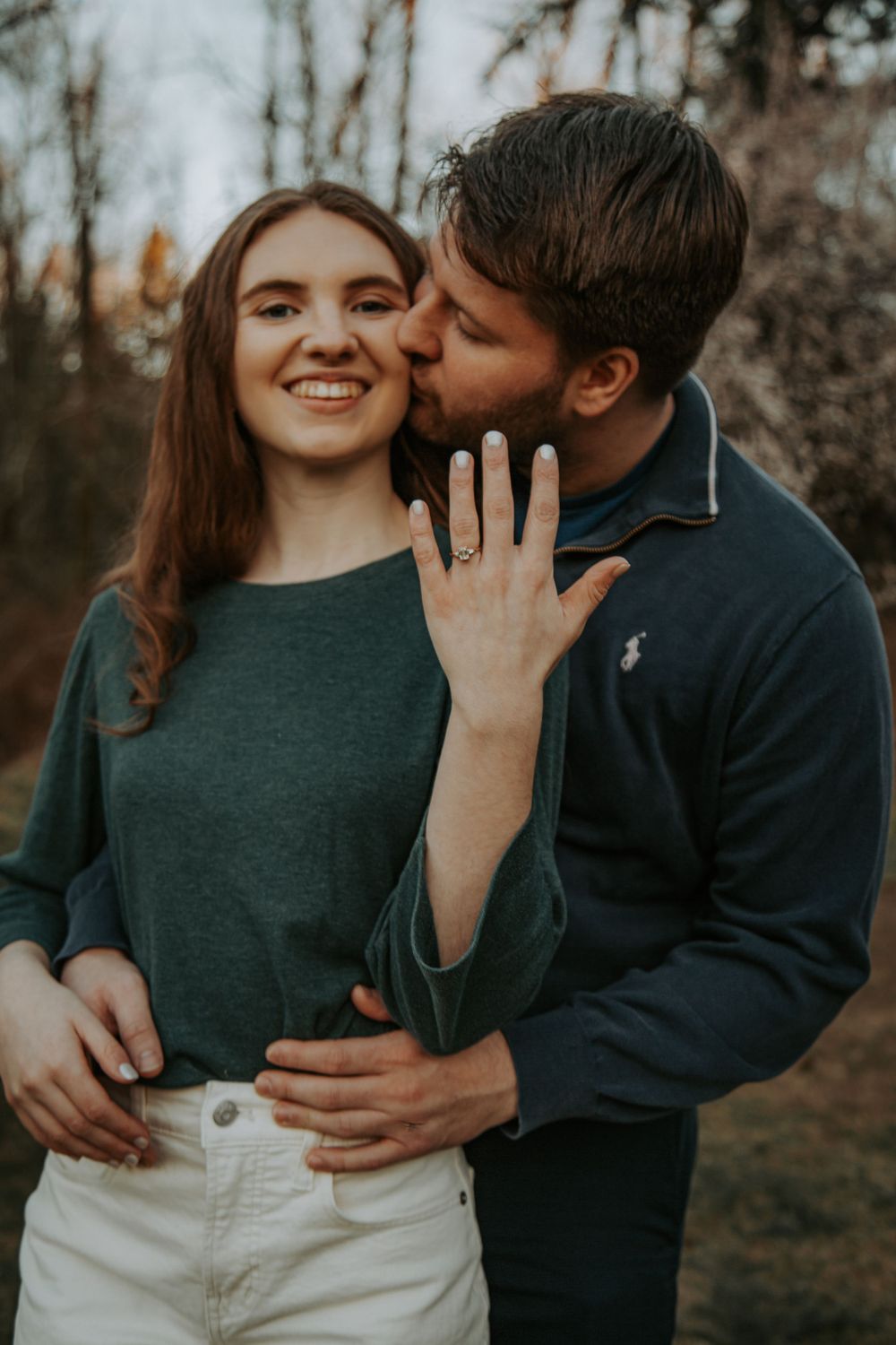A smiling woman shows her engagement ring to the camera while a man in a navy pullover kisses her cheek outdoors.