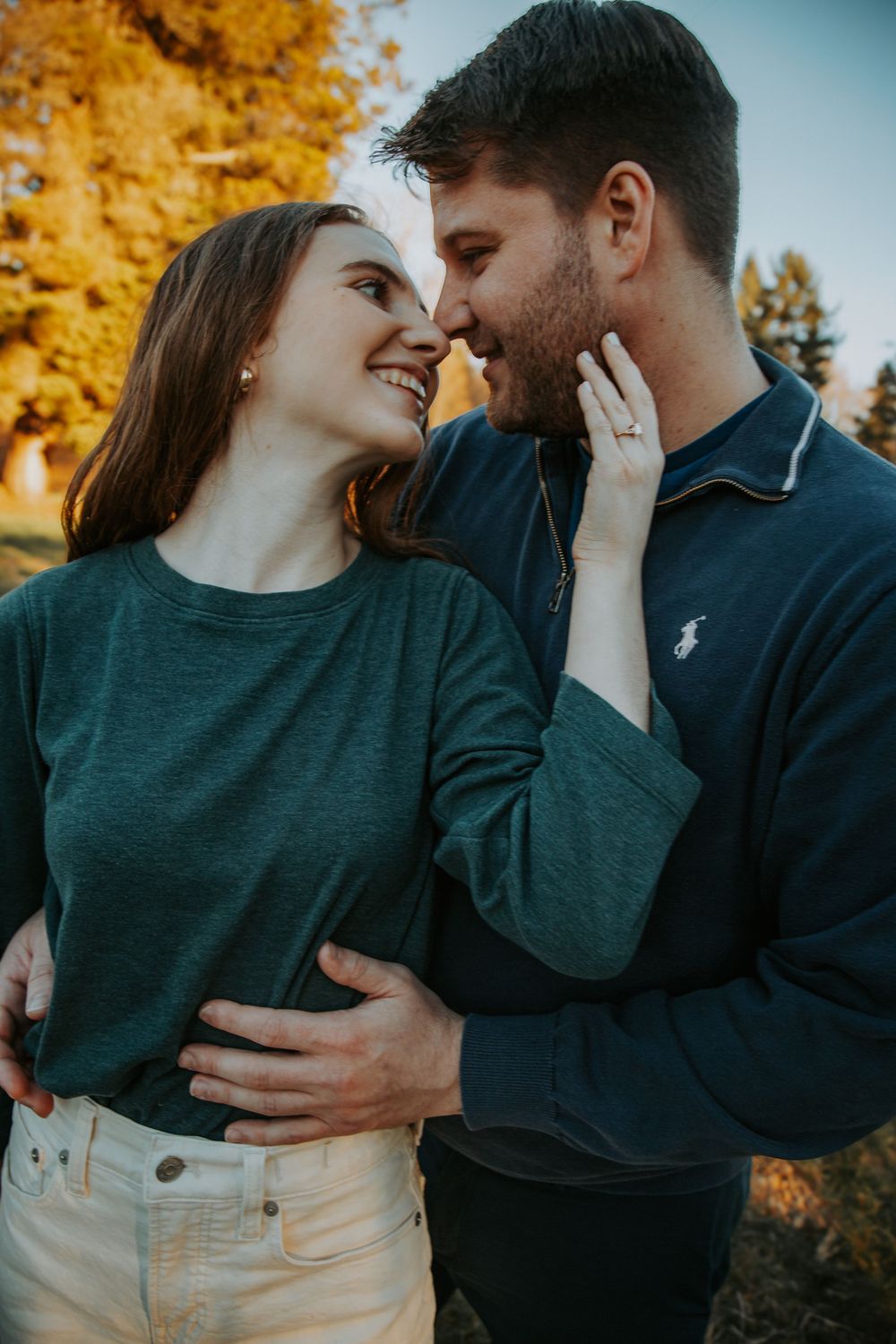 An engaged couple smiles at each other nose to nose in an autumn field as she shows her ring while he holds her waist.