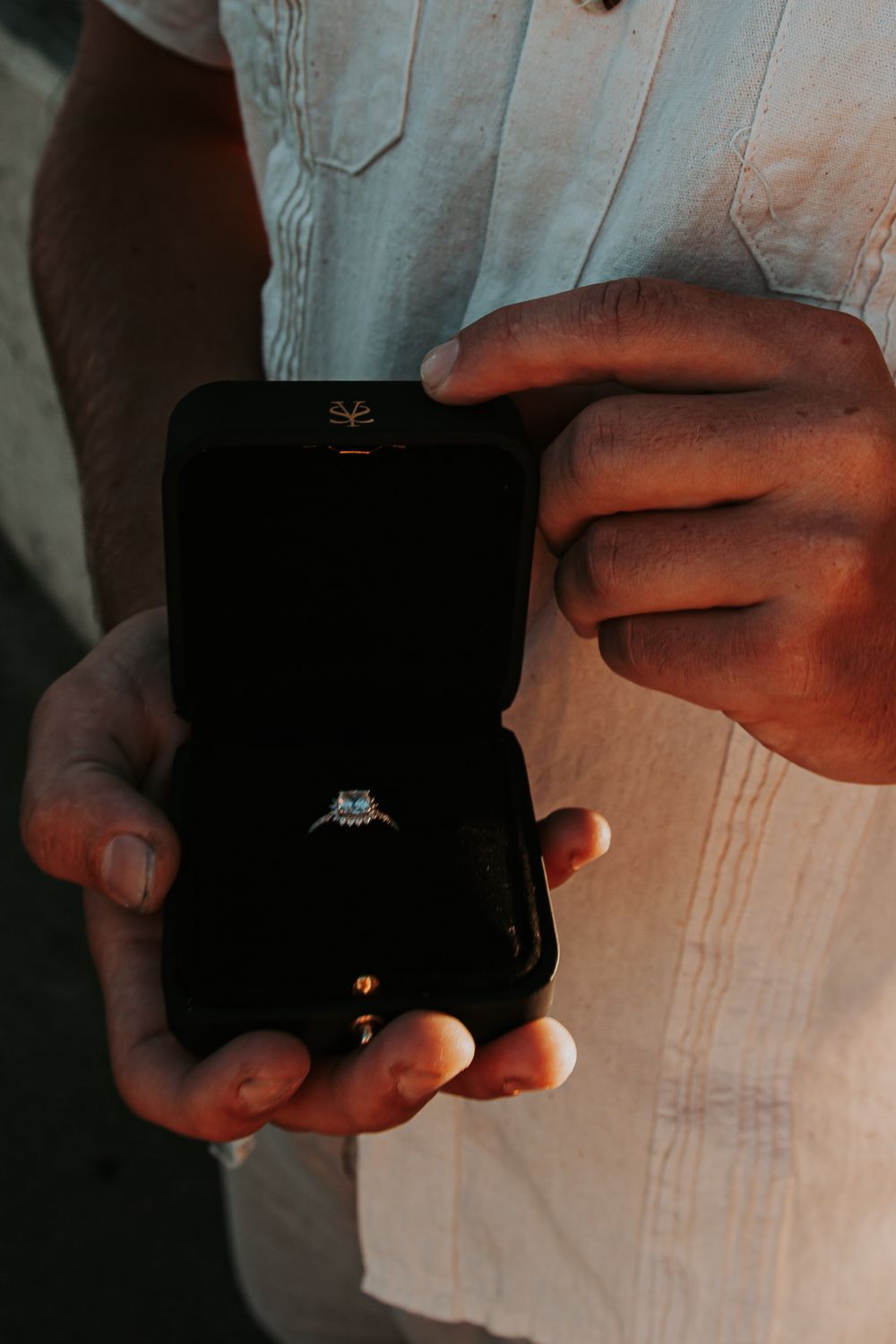 Hands hold an open black ring box displaying a diamond engagement ring, with a man in a white linen shirt behind.