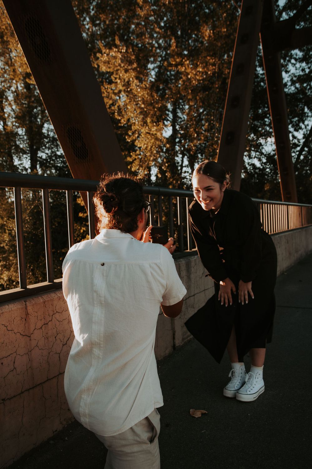 A smiling woman leans forward with hands clasped as a man kneels in front of her during a proposal on a bridge.