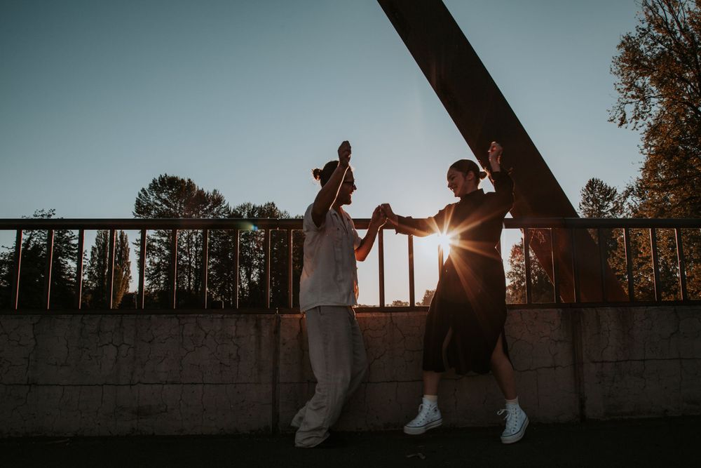 Two people hold hands and dance on a bridge walkway with the sun setting behind them and trees in the distance.