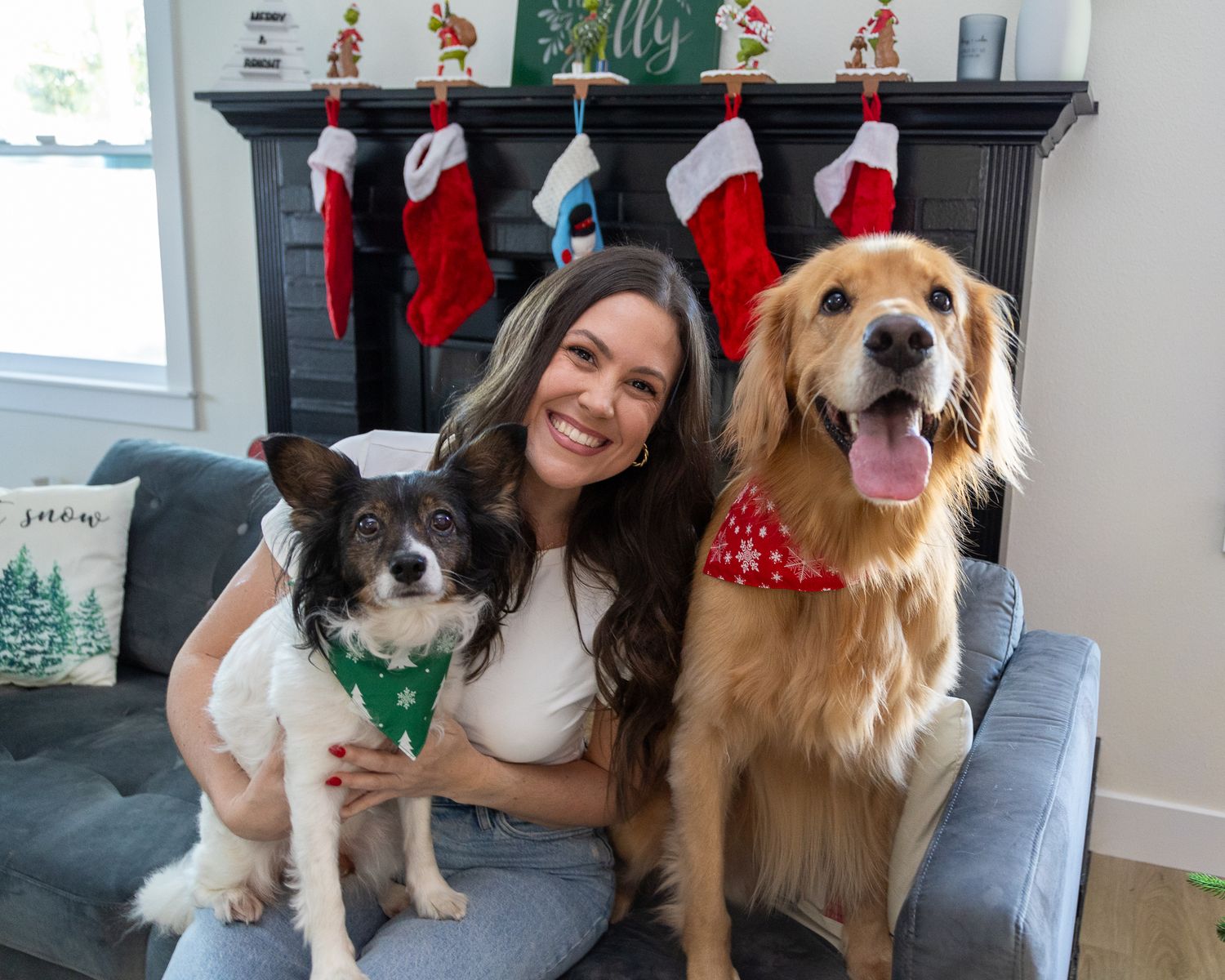 Brunette woman posing with two dogs in Christmas bandanas by a decorated fireplace.