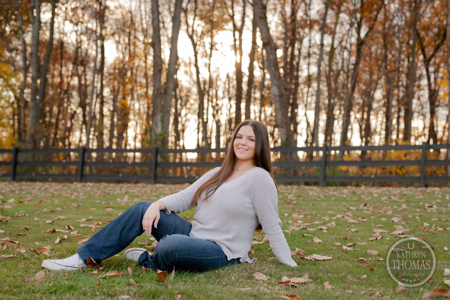 Relaxed outdoor portrait on grass with fall foliage and wooden fence backdrop at sunset.