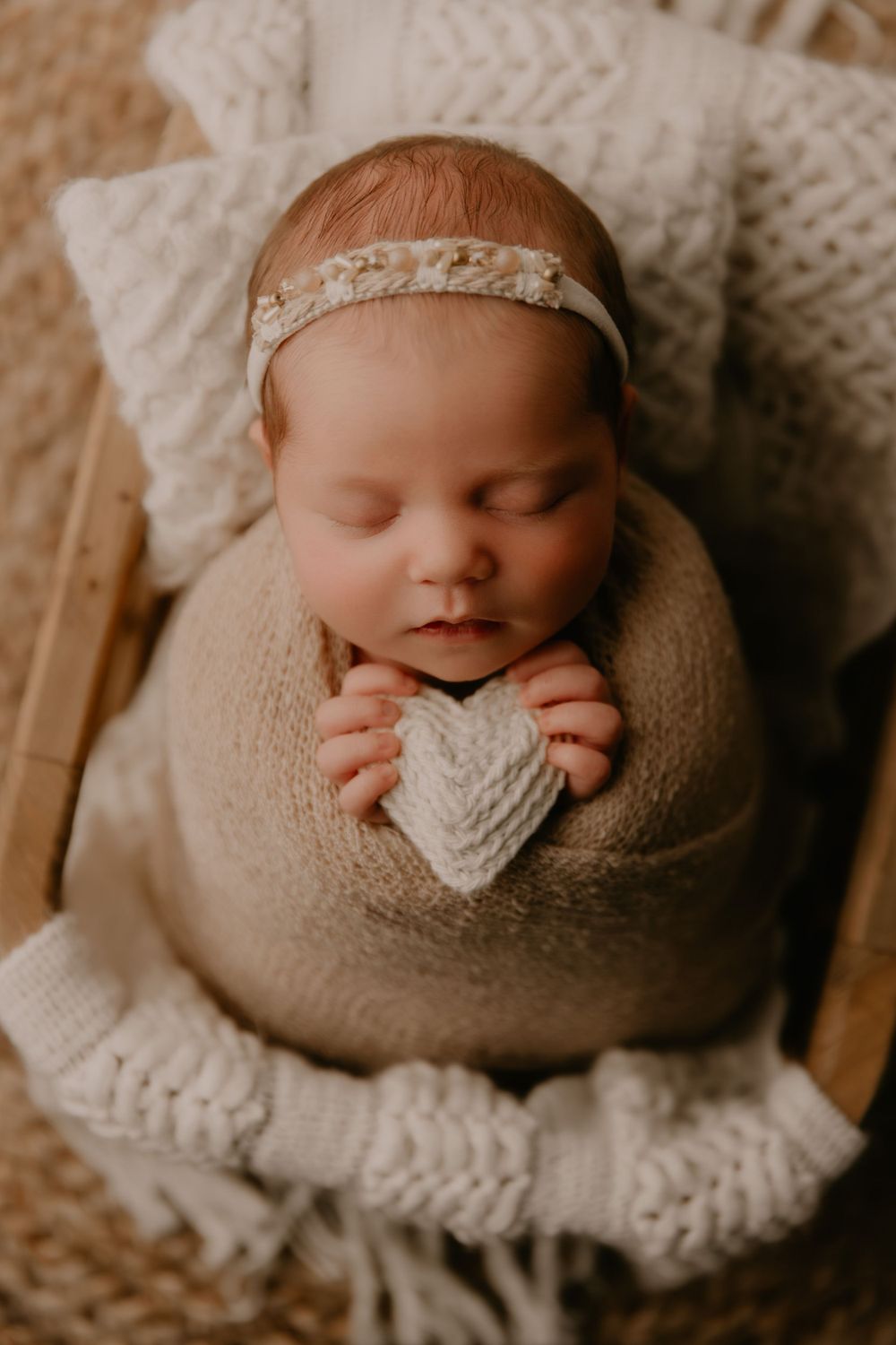 Peaceful sleeping newborn swaddled in beige wrap with lace headband resting on textured white blanket in warm lighting.
