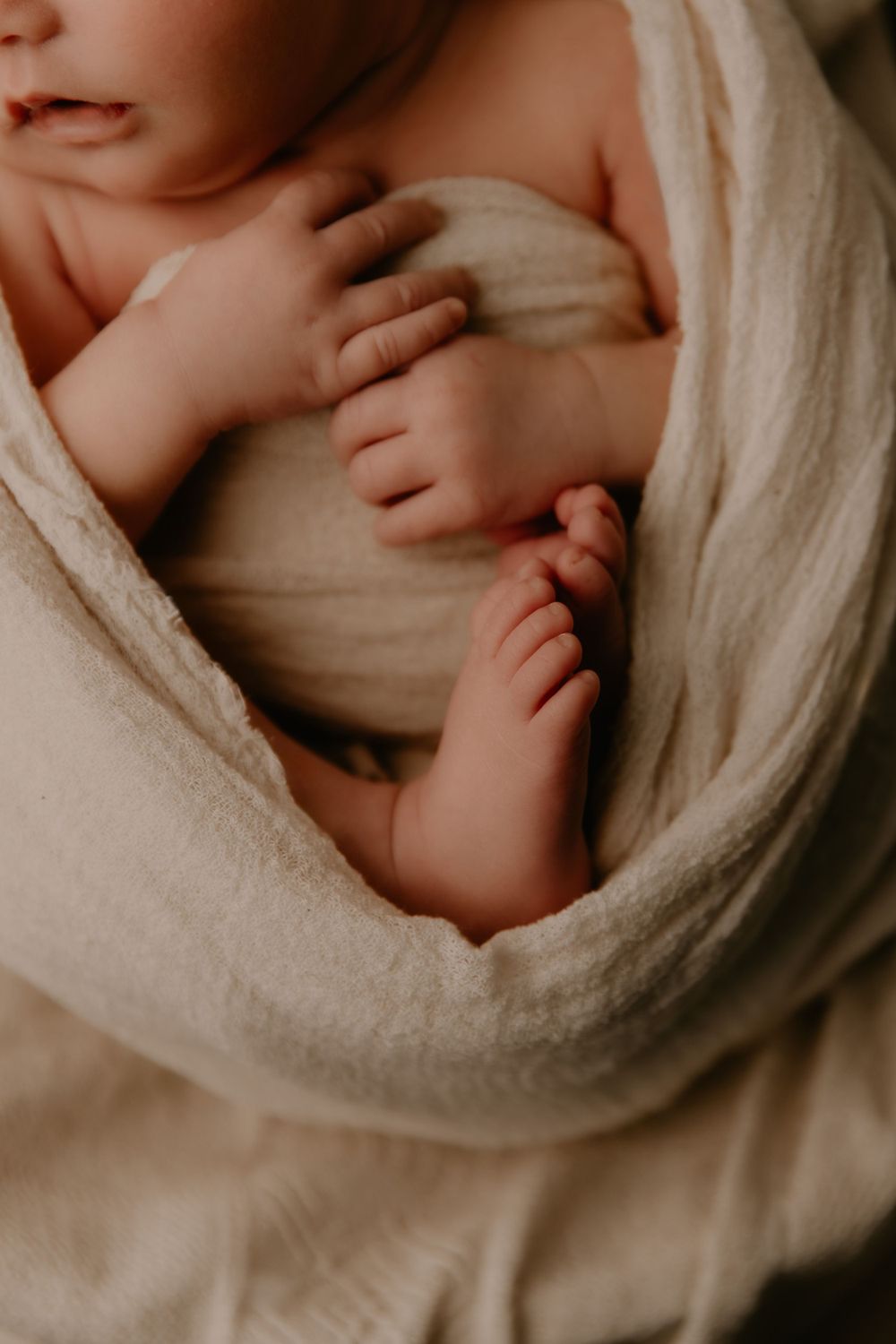 Close-up view of tiny baby hands and feet wrapped in soft beige towel with moody lighting.