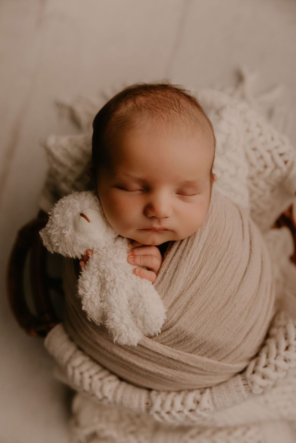 Peaceful sleeping newborn swaddled in cream fabric with white plush teddy bear.